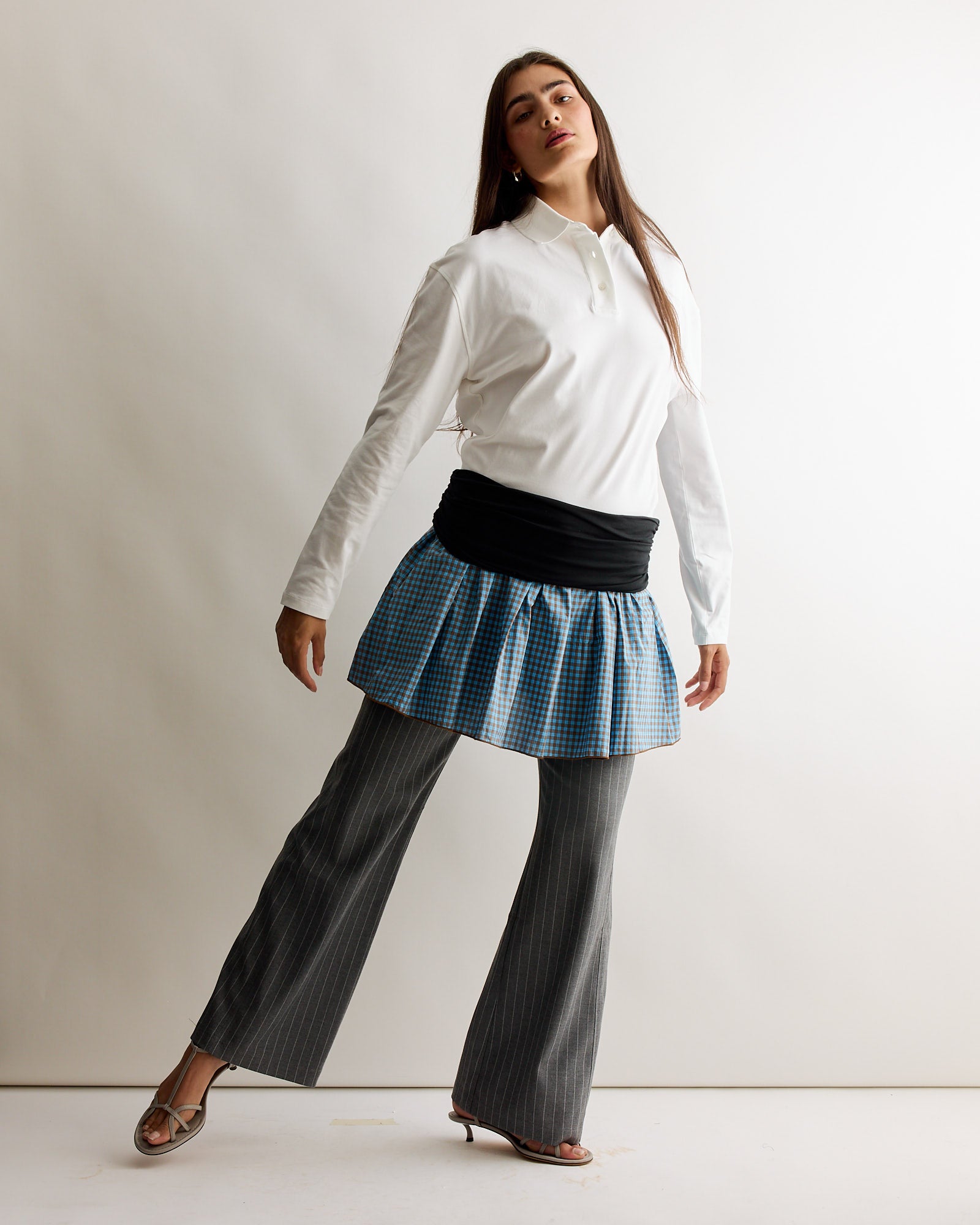 A woman poses confidently wearing Gimaguas' Tailored Nicole Trouser in Grey, styled beneath a blue plaid mini skirt, paired with a white long-sleeve shirt, a black waist wrap, and open-toed heels against a plain backdrop.