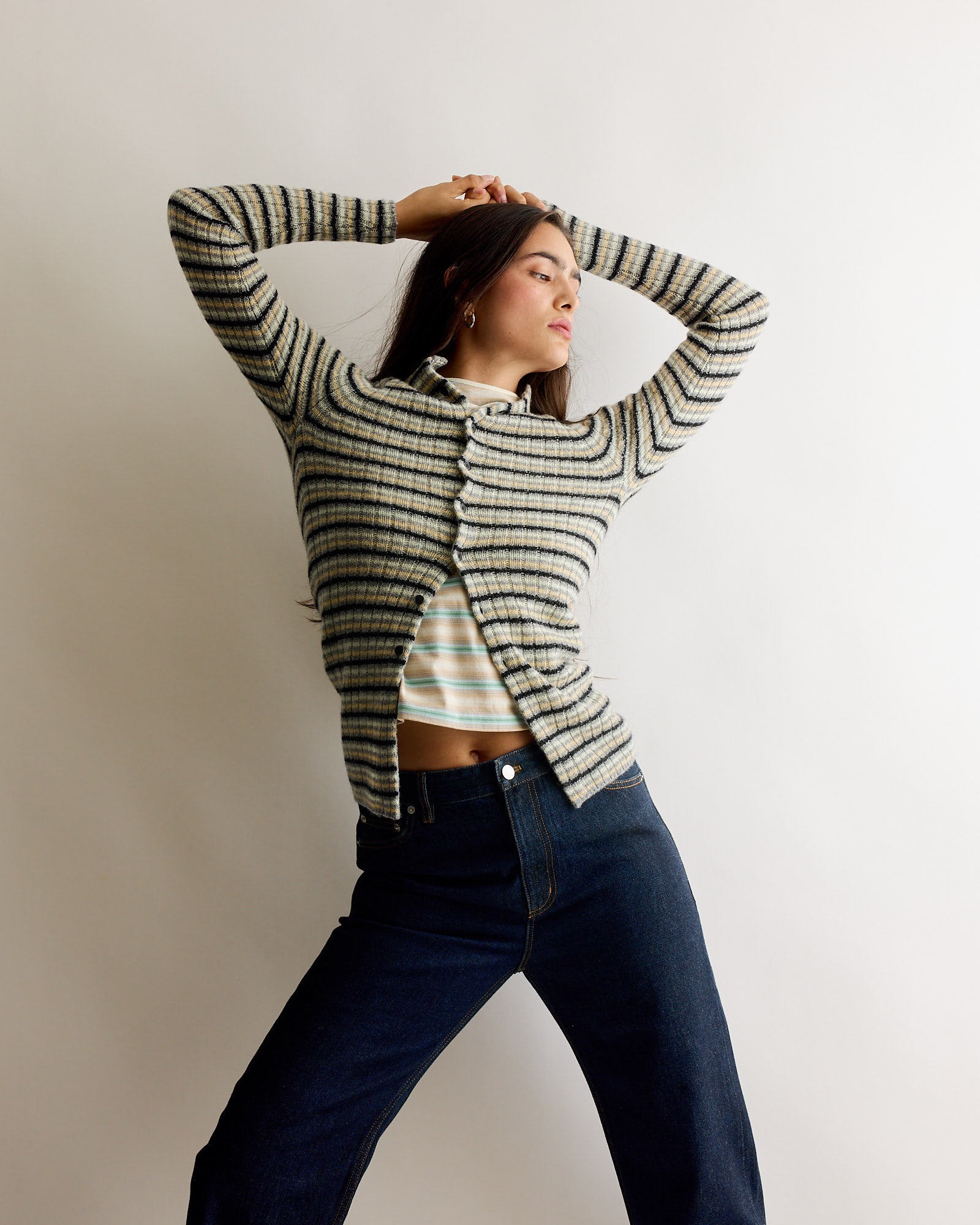 A woman poses confidently against a plain background, wearing the Auralee Gradient Jersey Tee in Fade Blue Stripe over a striped crop top and high-waisted dark jeans. With arms raised above her head, she gazes to the side.