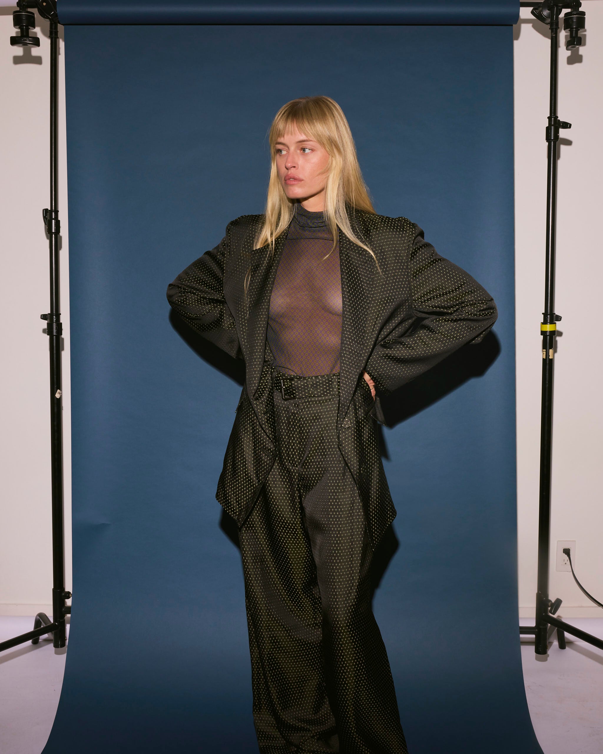 A woman with blonde hair stands before a blue backdrop, wearing Dries Van Noten's Belted Jacquard Trousers in Black over a sheer top, hands on hips and gazing to the side with a neutral expression.