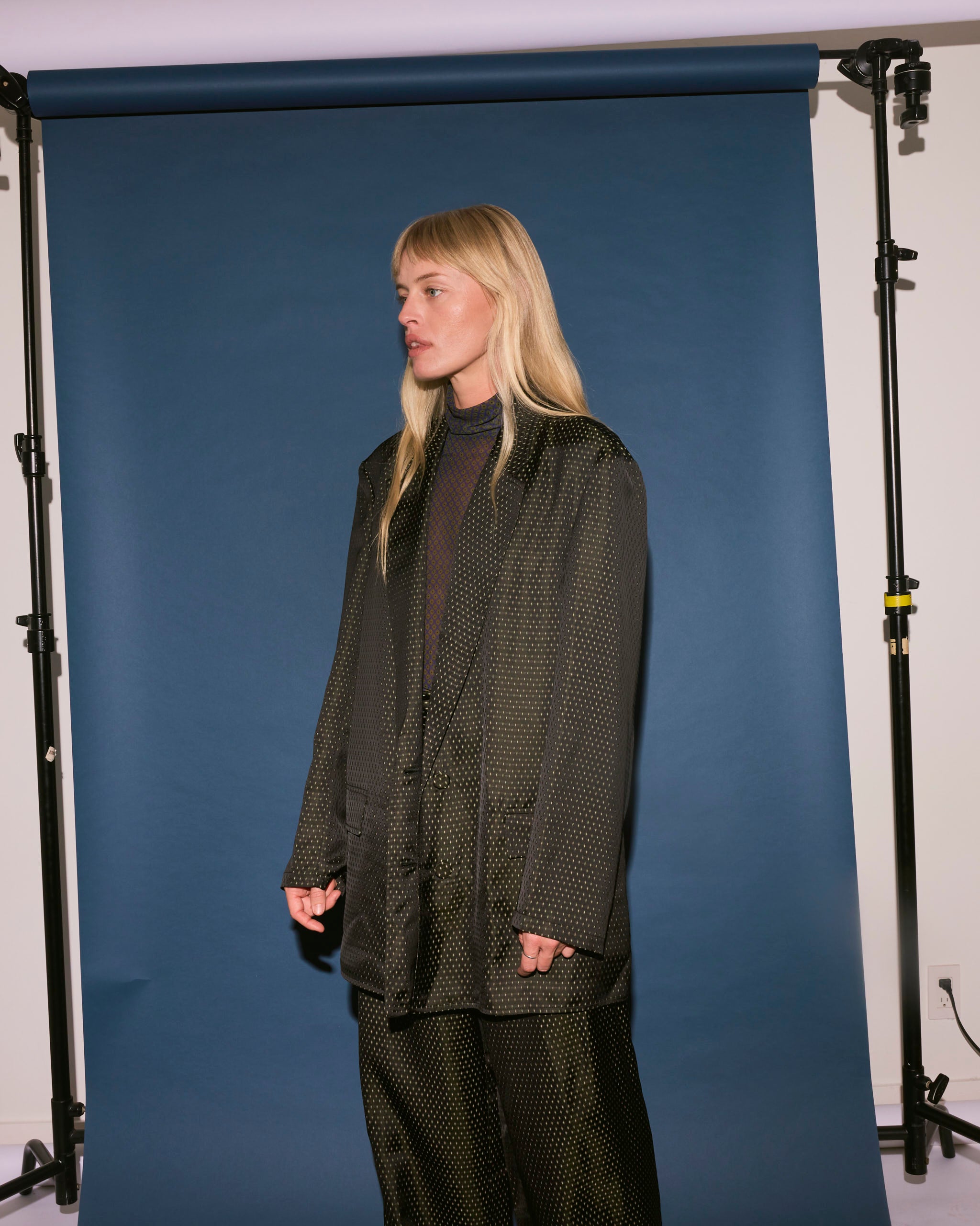 A person with long blonde hair wears the Dries Van Noten Jacquard Double-Breasted Blazer in Black, standing before a dark blue studio backdrop and looking to the left.