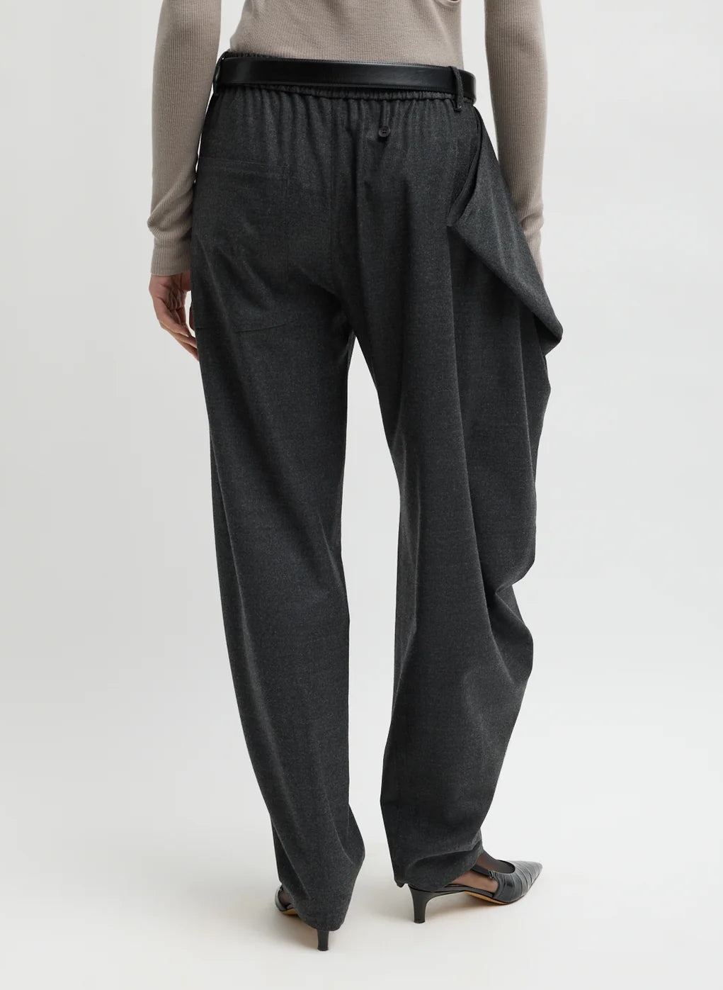 A person wearing the Tibi Cozy Flannel Side Drape Pant in Dark Heather Grey, featuring a loose fit and asymmetrical flap detail, styled with heels and shown from the back against a plain background.