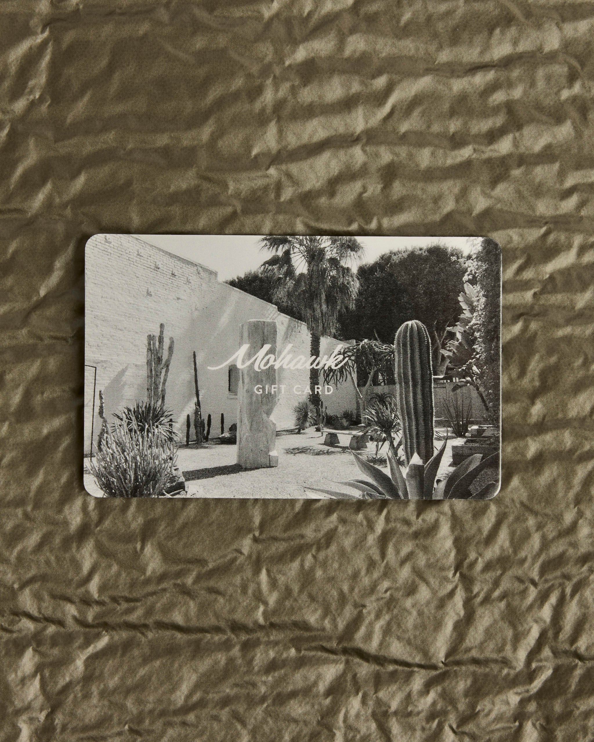 Give the perfect gift for any occasion with a Mohawk General Store E-Gift Card—featuring a black-and-white desert scene with cacti and a white building on a textured brown background.