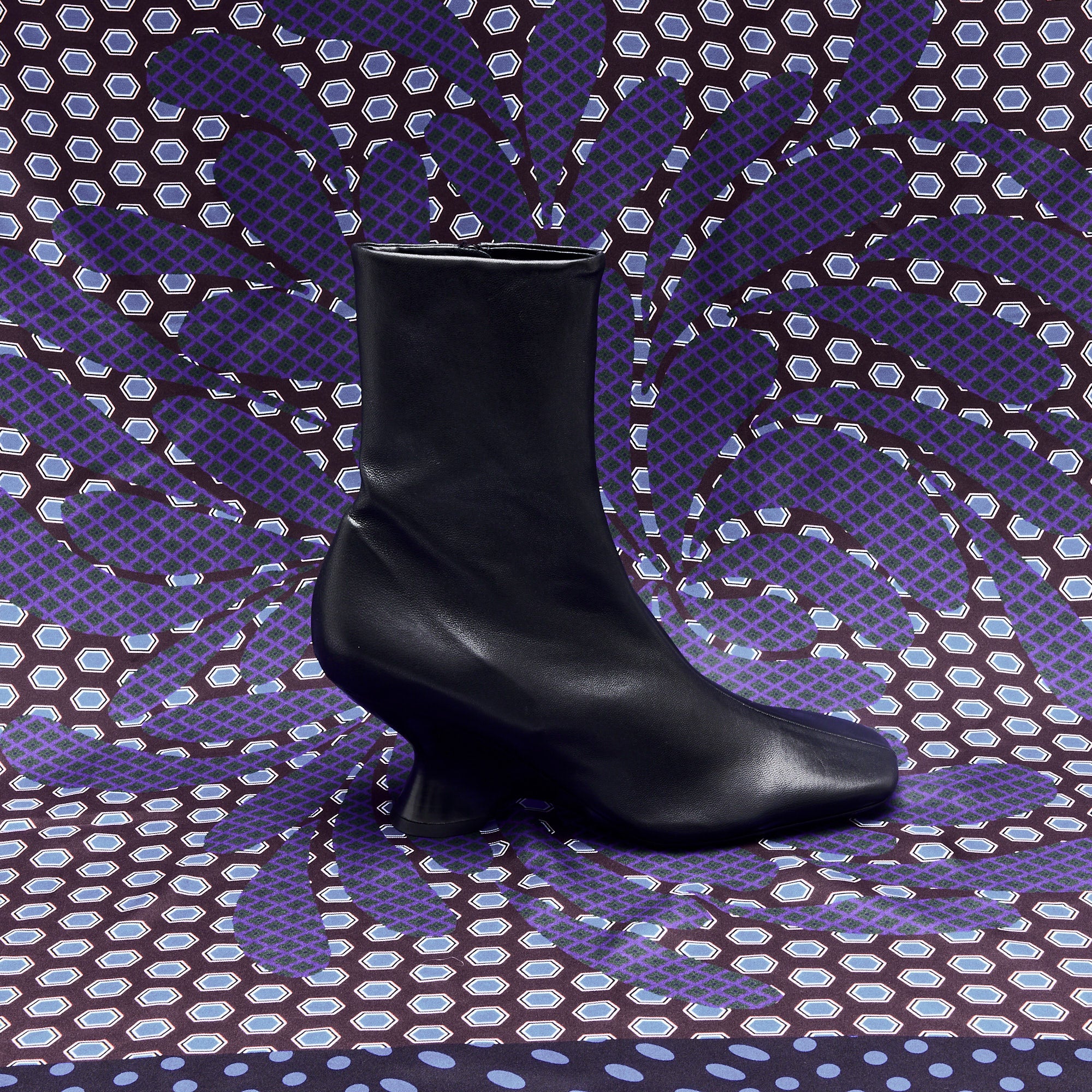 The Dries Van Noten Leather Wedge Boot in Black is displayed solo against a vibrant purple and blue patterned background with hexagonal and swirling shapes.