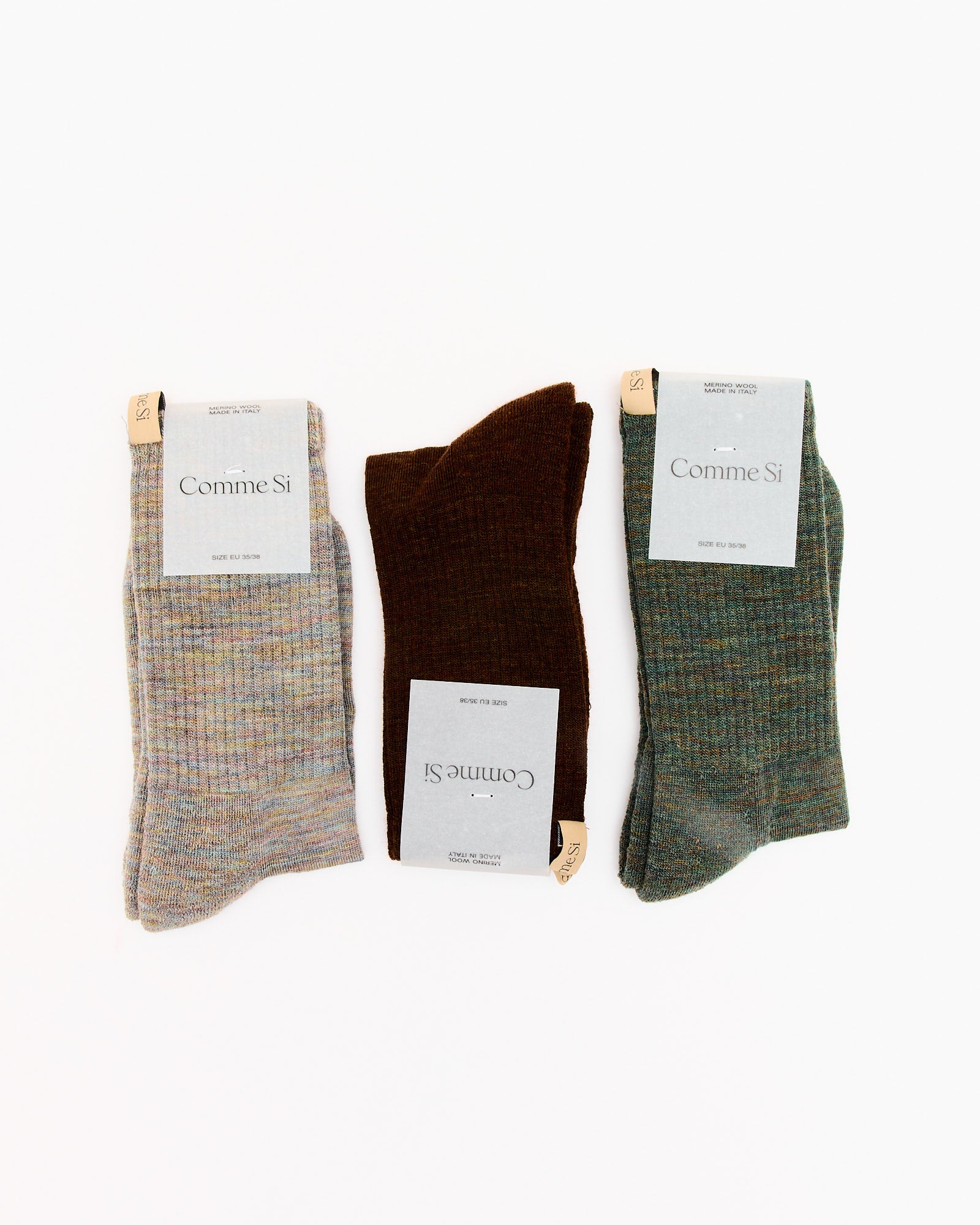 Three pairs of Comme Si's The Tube Sock in Cocoa are displayed folded on a white background: light gray, dark brown, and green. Each pair has a top label attached. For sizing questions, try our online chat.