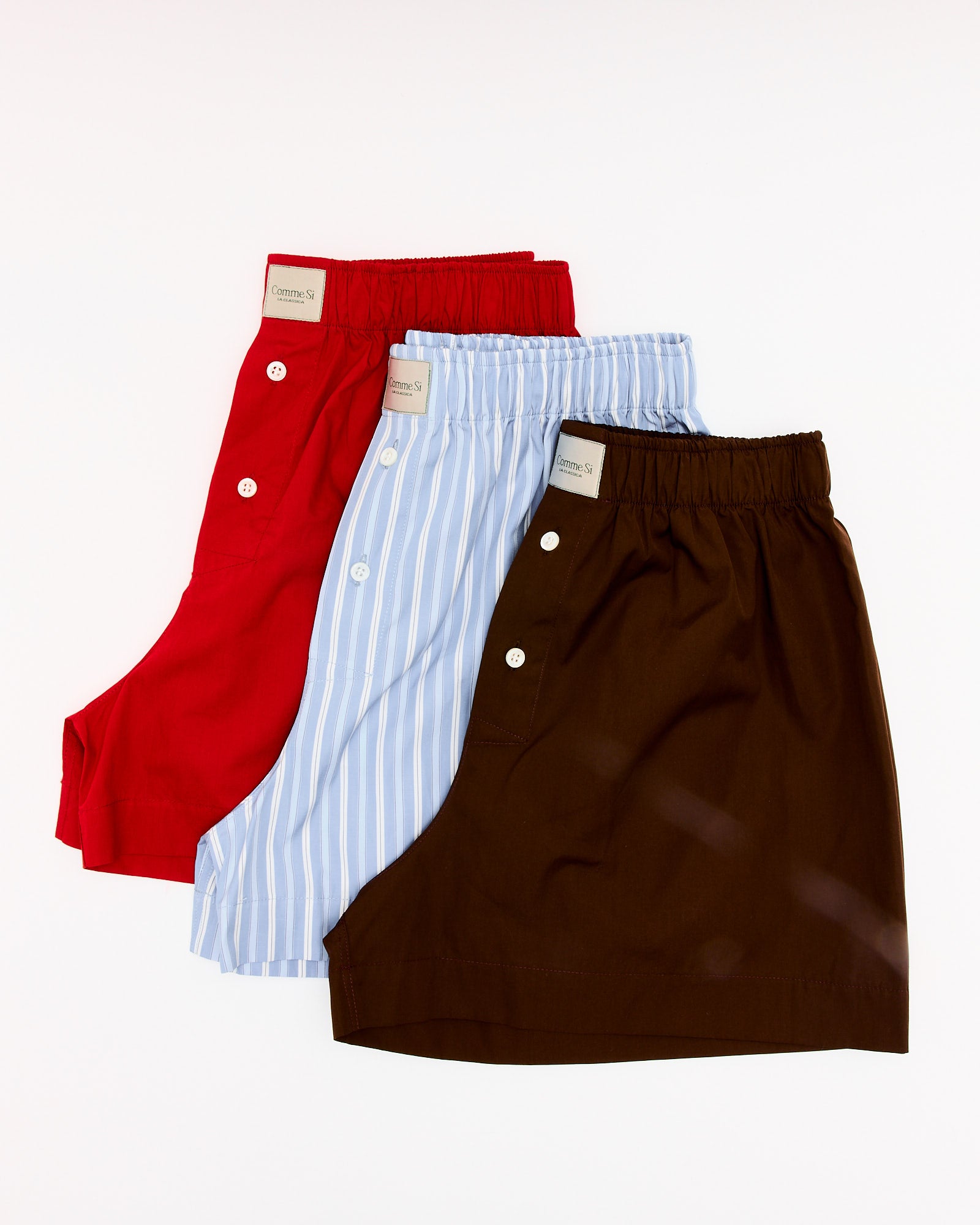 Three pairs of Comme Si La Boxer Classica in Maraschino are displayed in red, light blue with white stripes, and brown. Each Italian cotton boxer features a front button and branded waistband label—ideal for luxury loungewear.