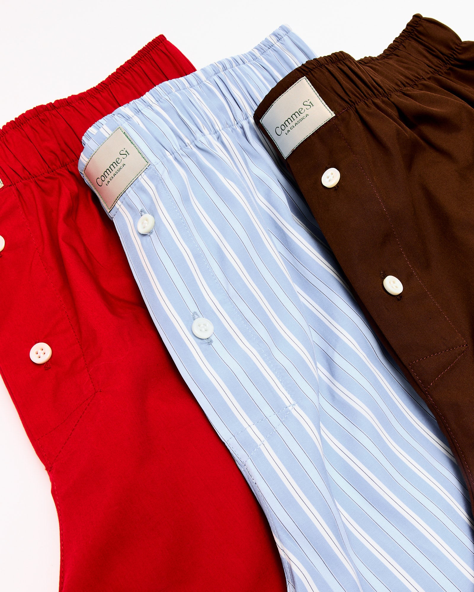 Three pairs of Comme Si La Boxer Classica in Executive Stripe shorts—each featuring a button fly and branded waistband—are shown in solid red, light blue with white stripes, and solid brown. For sizing help, contact us via online chat.