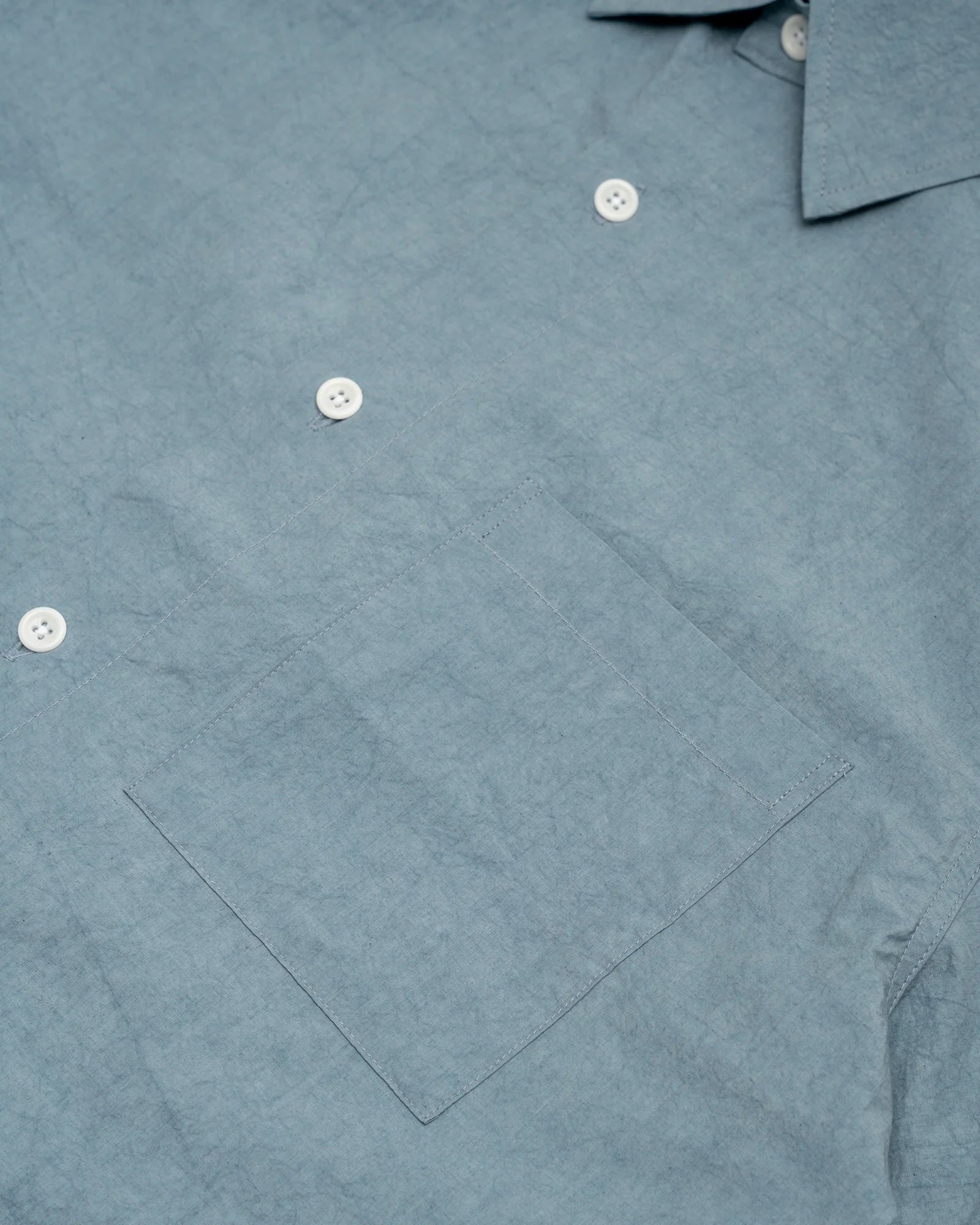 Close-up of the James Coward Short Sleeve Shirt in Blue, made in Vancouver. Features include white buttons, a left chest pocket, part of the collar, and slightly wrinkled Japanese typewriter cloth fabric.
