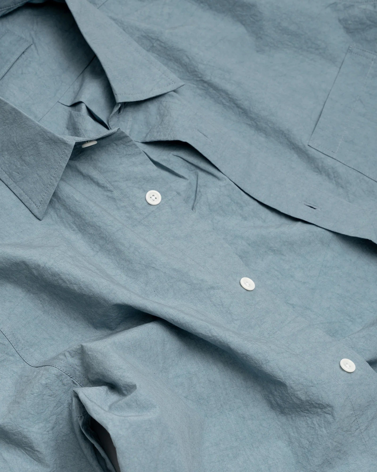 A close-up of the James Coward Short Sleeve Shirt in Blue, featuring crinkled Japanese typewriter cloth, white buttons, a pointed collar, and a visible chest pocket, laid flat.