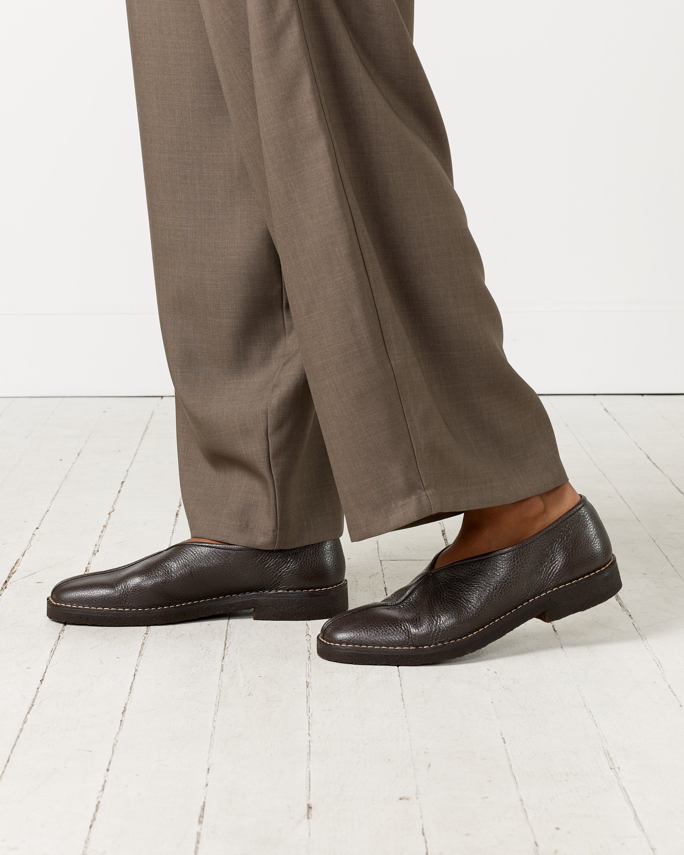 A person wearing loose-fitting brown trousers and Lemaire’s Piped Crepe Slipper in Dark Brown stands on a white wooden floor, with only their lower legs and feet visible to highlight the sleek grained leather of the shoes.