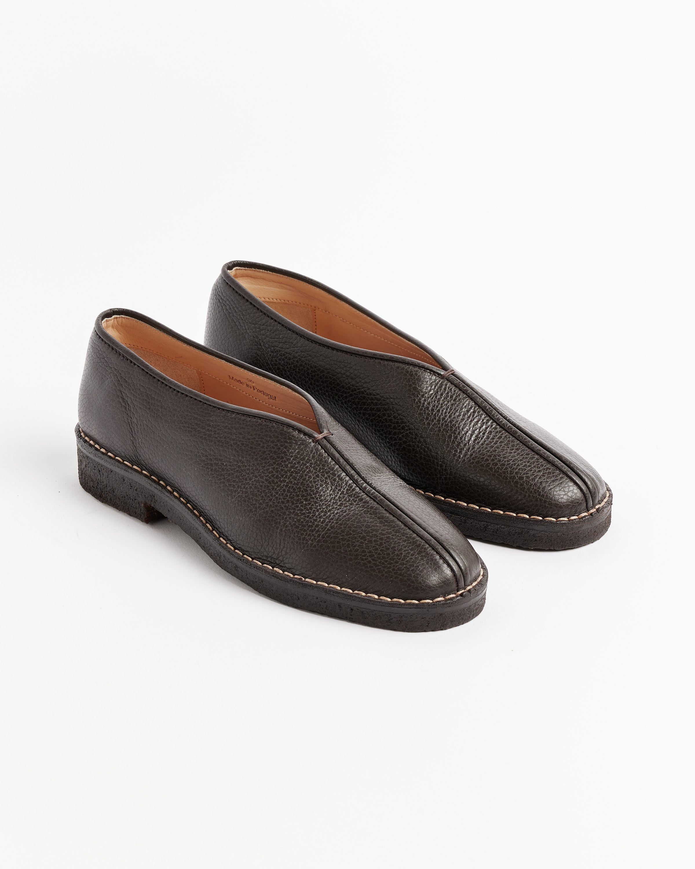 The Lemaire Piped Crepe Slipper in dark brown features grained leather, a rounded toe, visible stitching along the sole, and a low heel, shown on a white background.