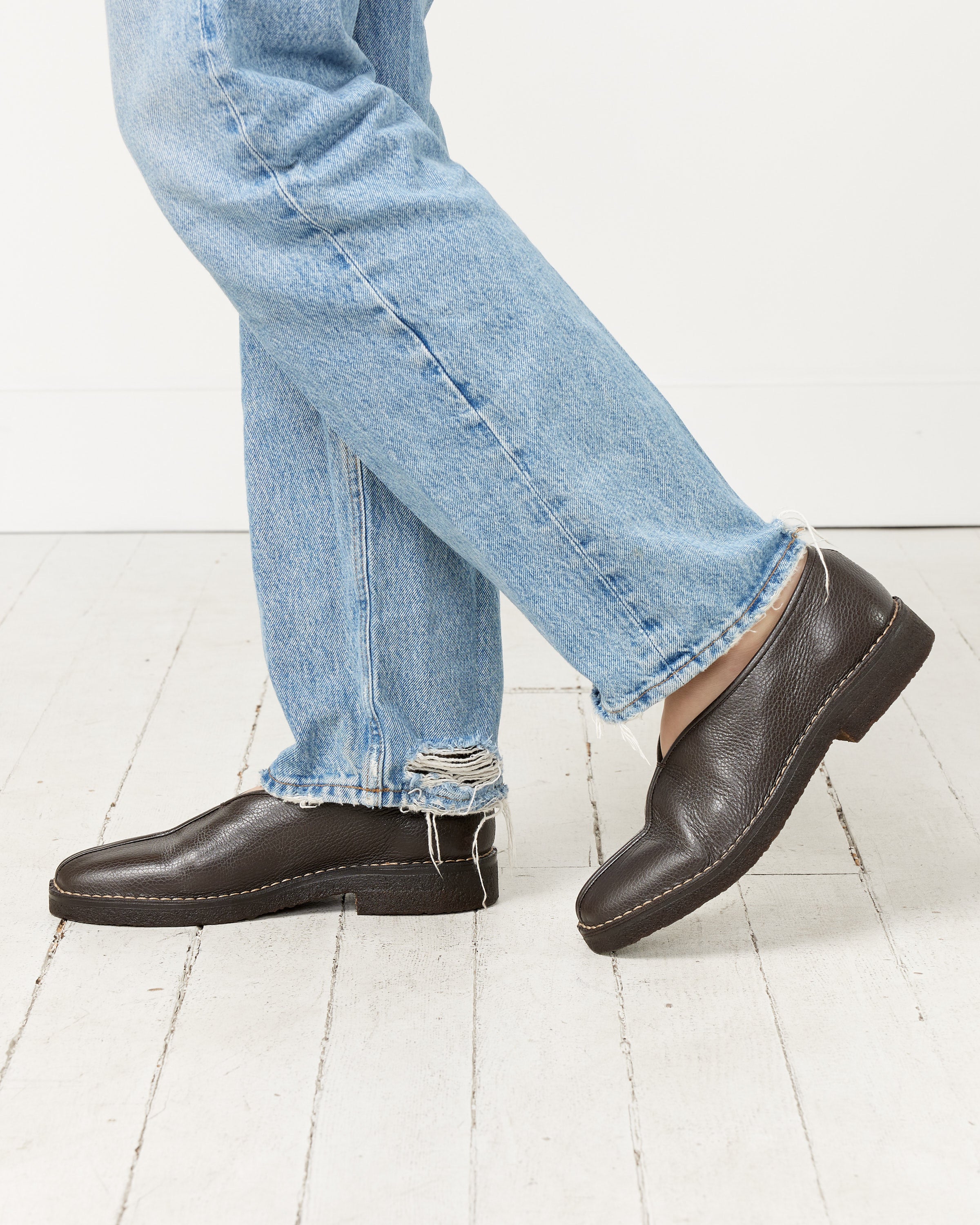 A person stands on a white wooden floor, one foot crossed over the other, wearing light blue, slightly distressed jeans and Lemaire’s Piped Crepe Slipper in Dark Brown.