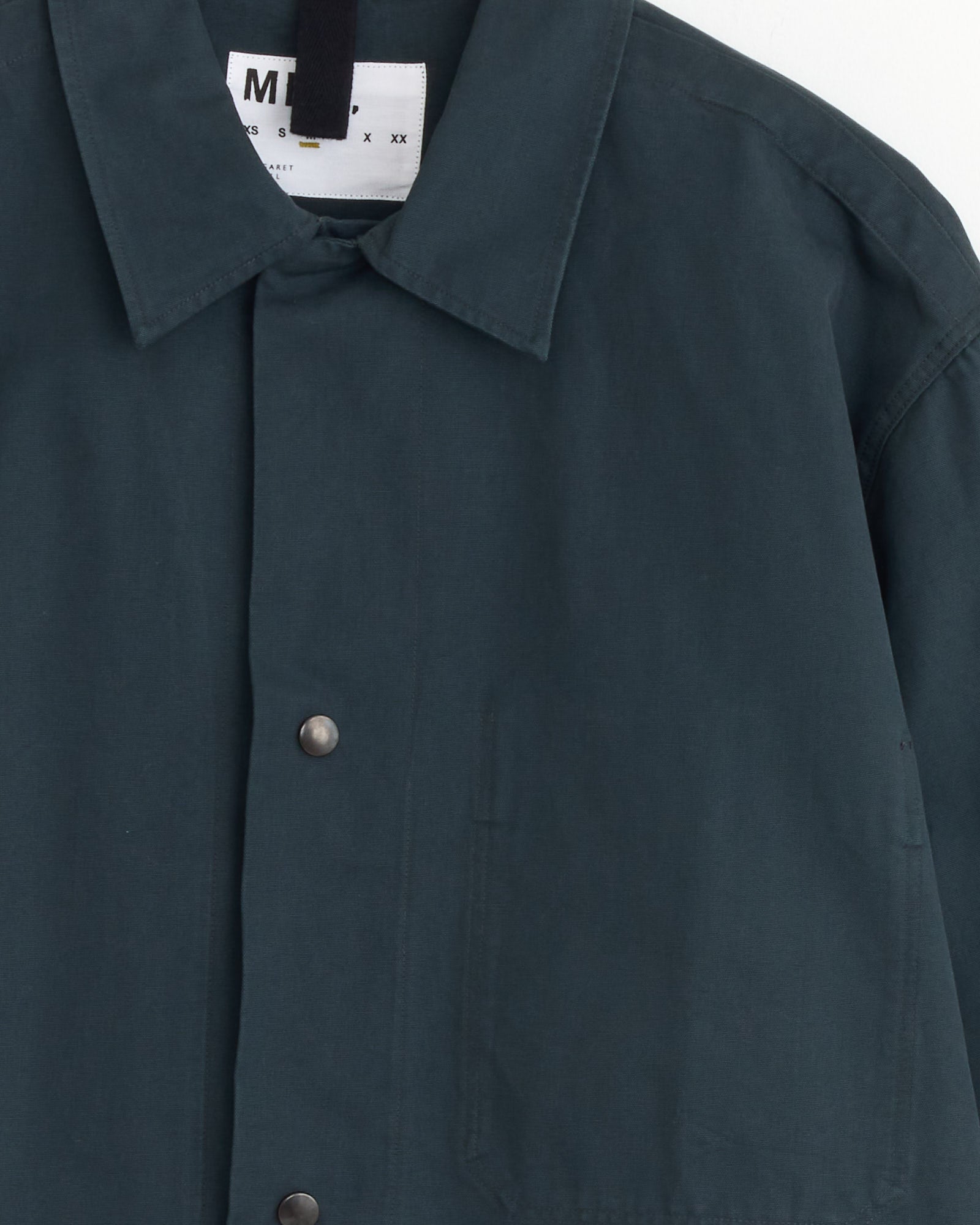 A close-up of the MHL by Margaret Howell Chore Shirt in Petrol shows its collar, metal snap buttons, and chest pocket, with a white branded label partially visible inside—ideal for checking fit or sizing before purchase.
