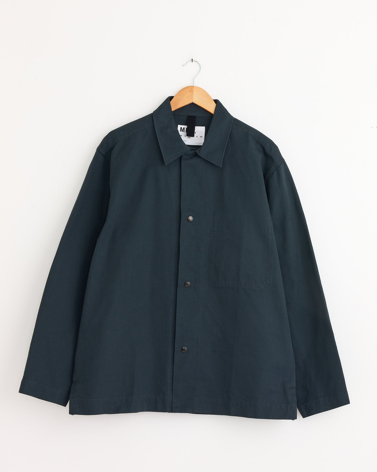 The MHL by Margaret Howell Chore Shirt in Petrol features a collar, chest pocket, and long sleeves. Shown here on a wooden hanger against a white wall. For fit or sizing help, contact us via online chat.