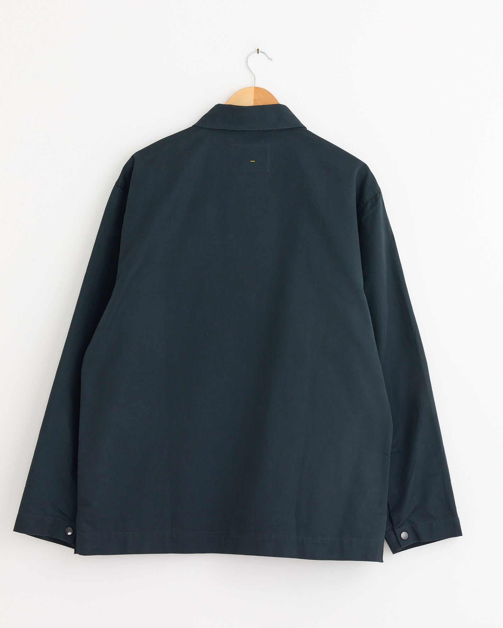 The Chore Shirt in Petrol by MHL by Margaret Howell, a dark long-sleeve button-up, hangs on a wooden hanger against a plain white wall, shown from the back. For sizing and fit details, contact us via online chat before ordering.