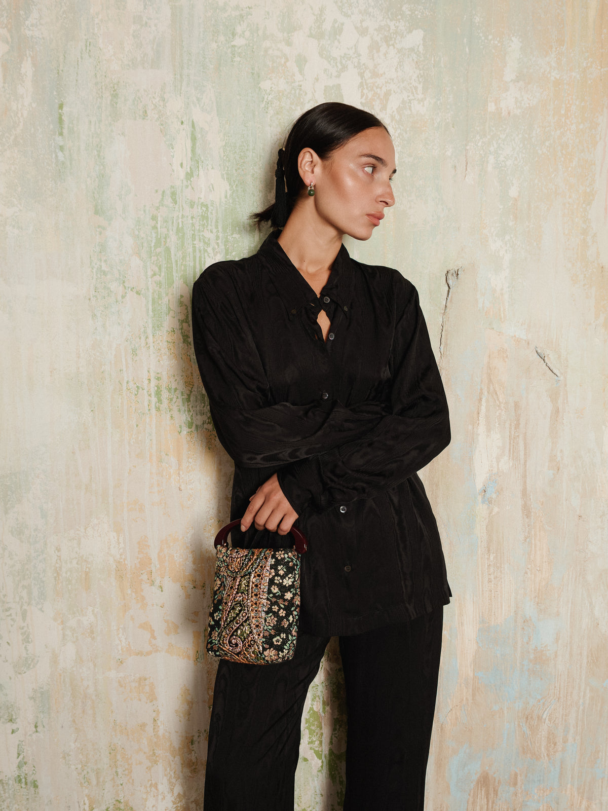 A woman in a black outfit stands against a textured wall, looking to the side with her arms crossed, holding the Dries Van Noten Embellished Bag in Brown.