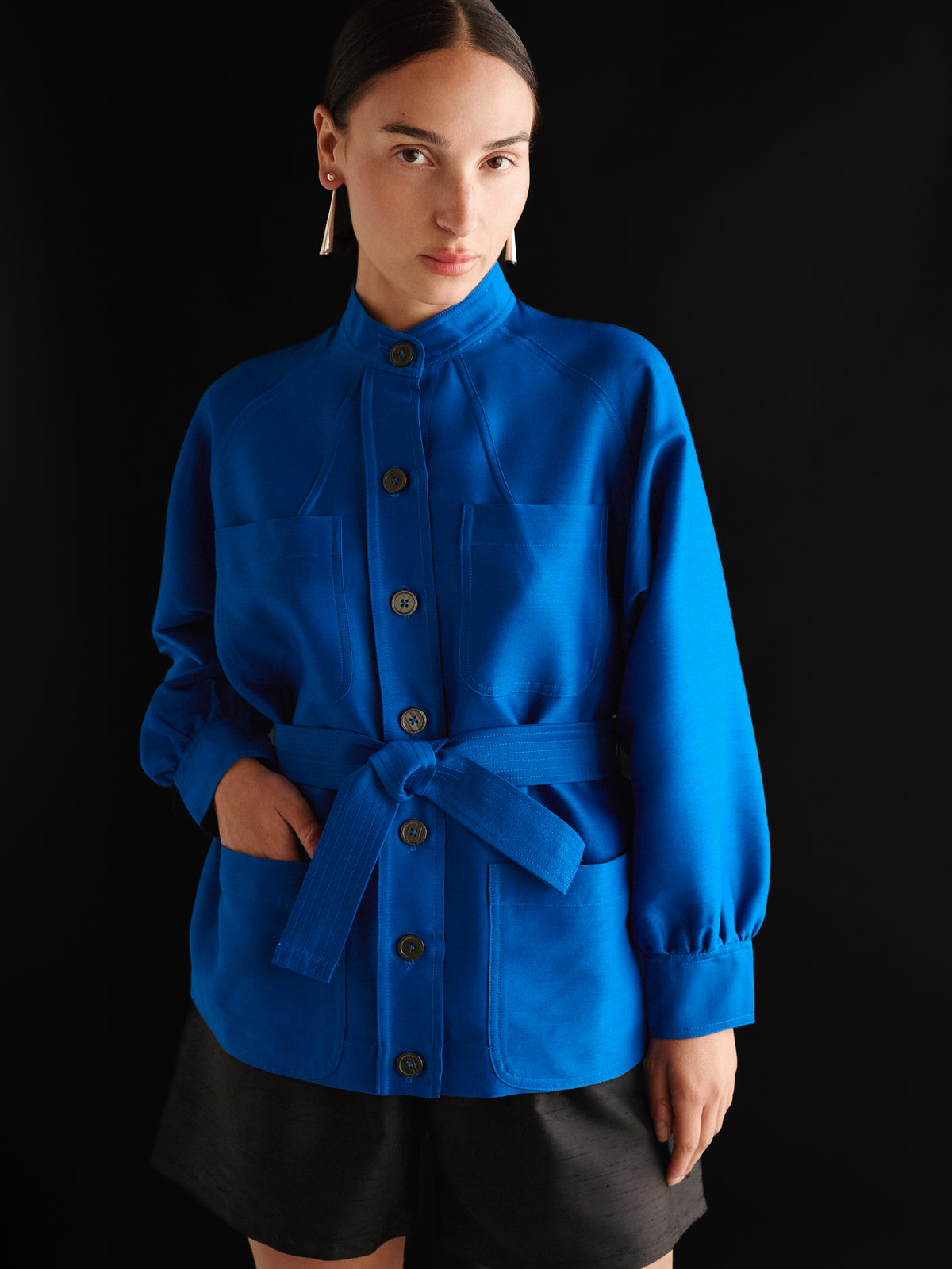 A woman wearing the Chelsea Mak Kiwa Jacket in Yves Blue, featuring a belted button-up design, and black shorts stands against a black background, looking at the camera.