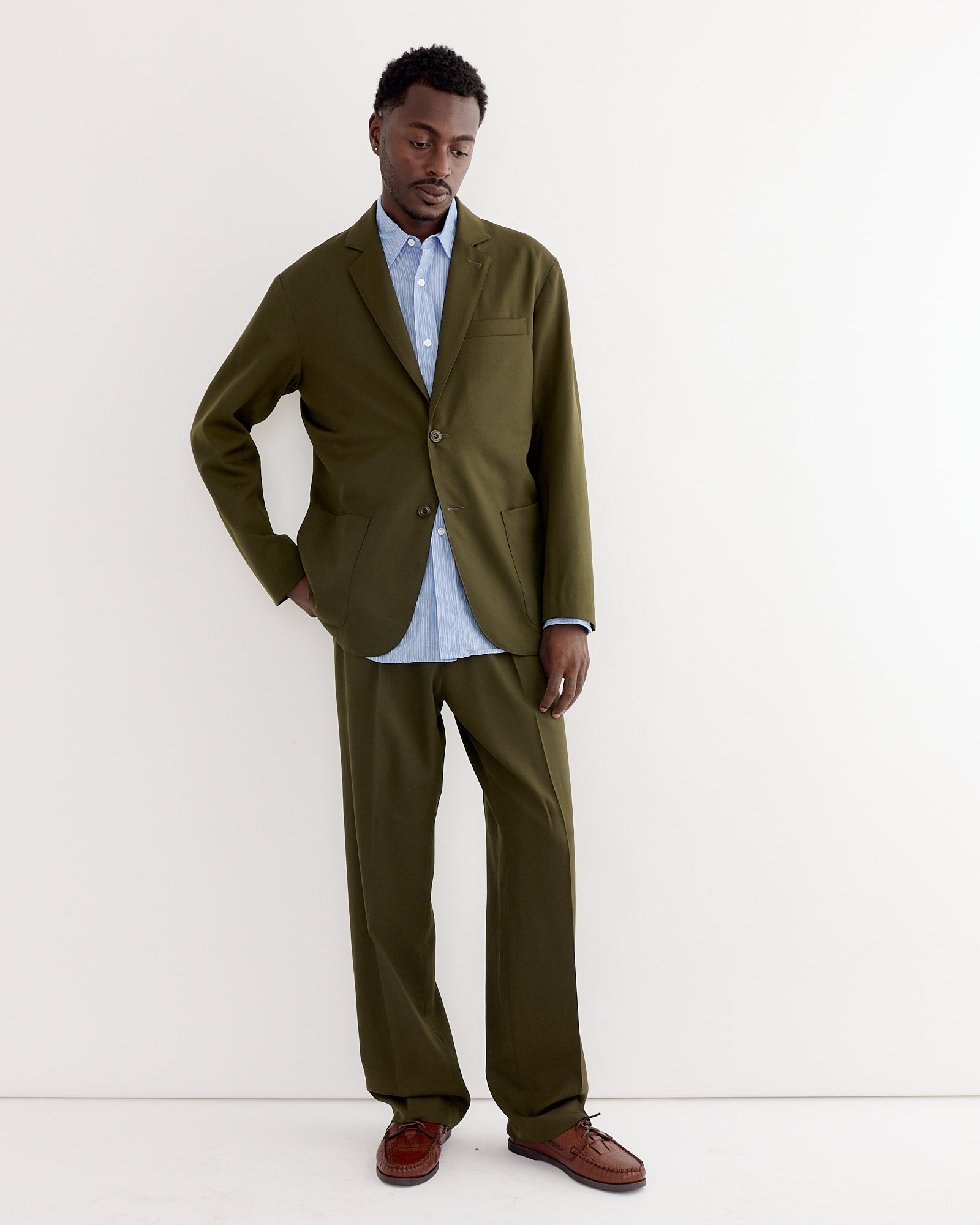 A man stands against a white wall wearing the SMOCK Blazer in Wool Gabardine Moss, paired with a light blue button-up shirt and brown dress shoes. One hand is in his pocket as he looks slightly downward with a relaxed expression.