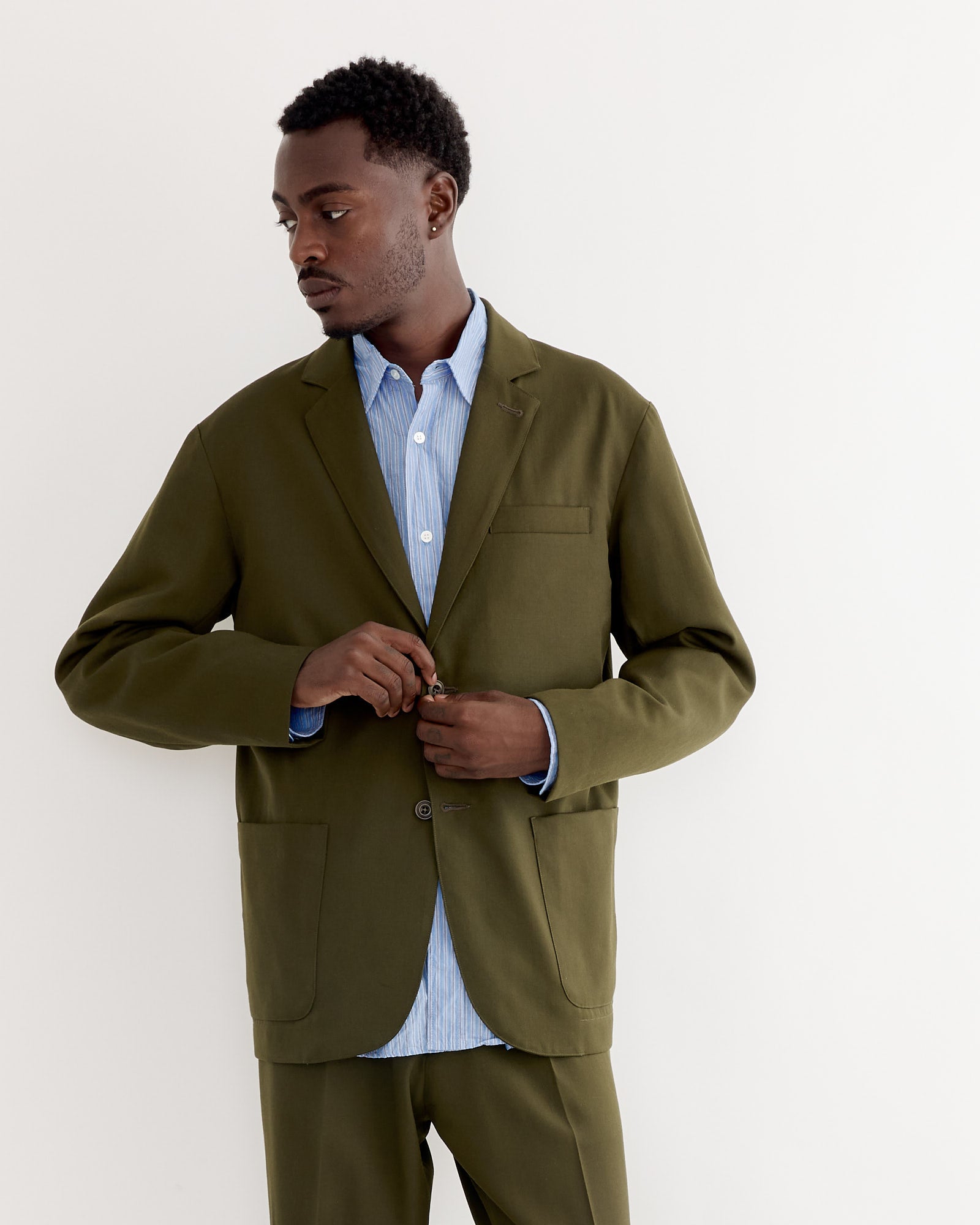 A man stands against a plain white background, wearing the SMOCK Blazer in Wool Gabardine Moss over a light blue button-up shirt. He looks to the side while buttoning his jacket.