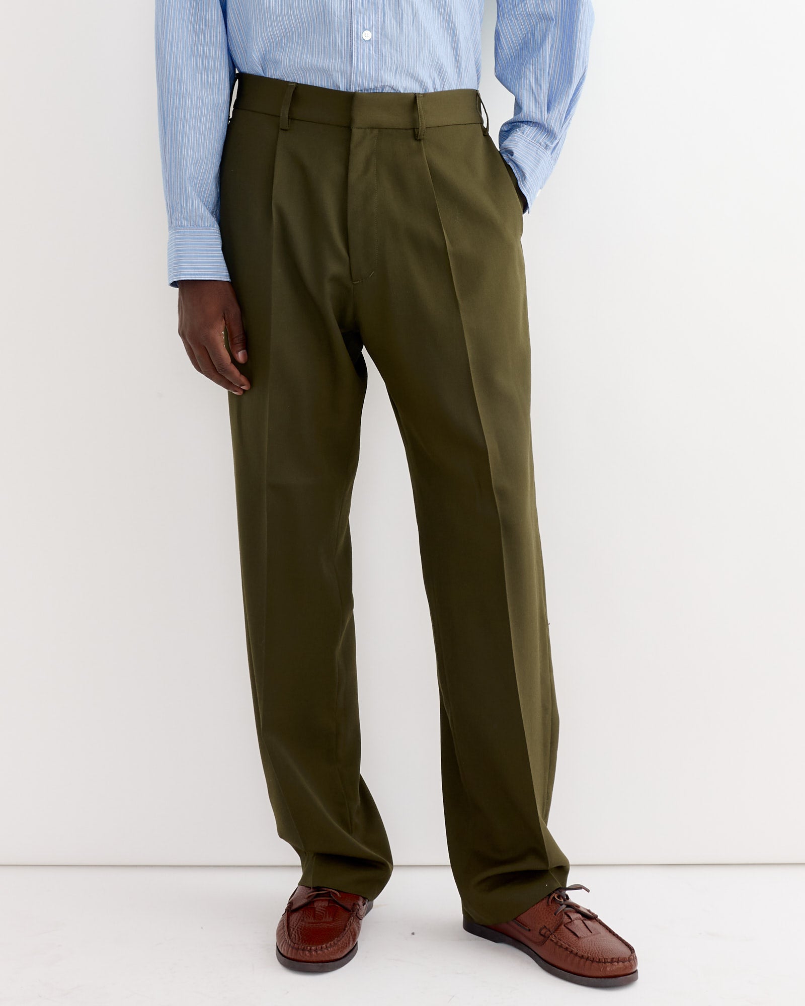 A person models the SMOCK West Coast Pant in Wool Gabardine Moss, paired with a light blue striped shirt and brown dress shoes, standing against a plain white background with their left hand in their pocket.