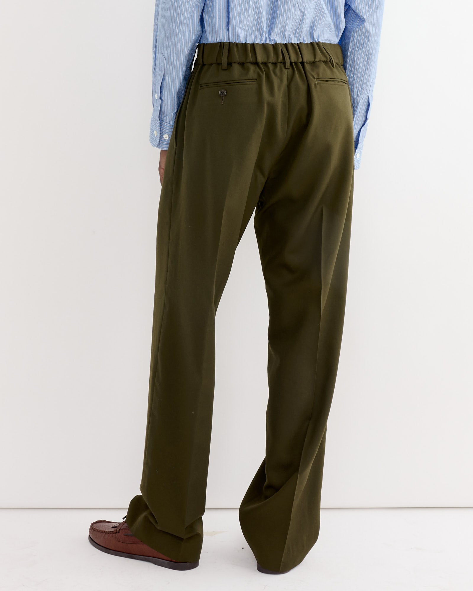 A person stands against a white wall, seen from behind, wearing SMOCK's West Coast Pant in Wool Gabardine Moss styled as loose-fitting dress pants, paired with brown loafers and a blue striped button-up shirt with sleeves rolled down.