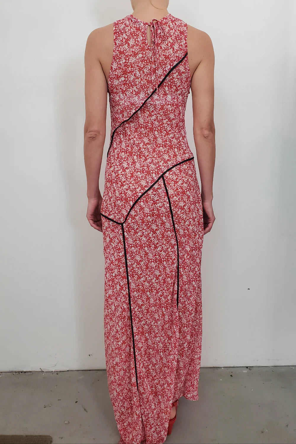 A person stands facing away, wearing the NOMIA Paneled Bias Maxi Dress in Poppy Floral with black diagonal trim, an asymmetrical hem and design, and a small upper back keyhole opening.