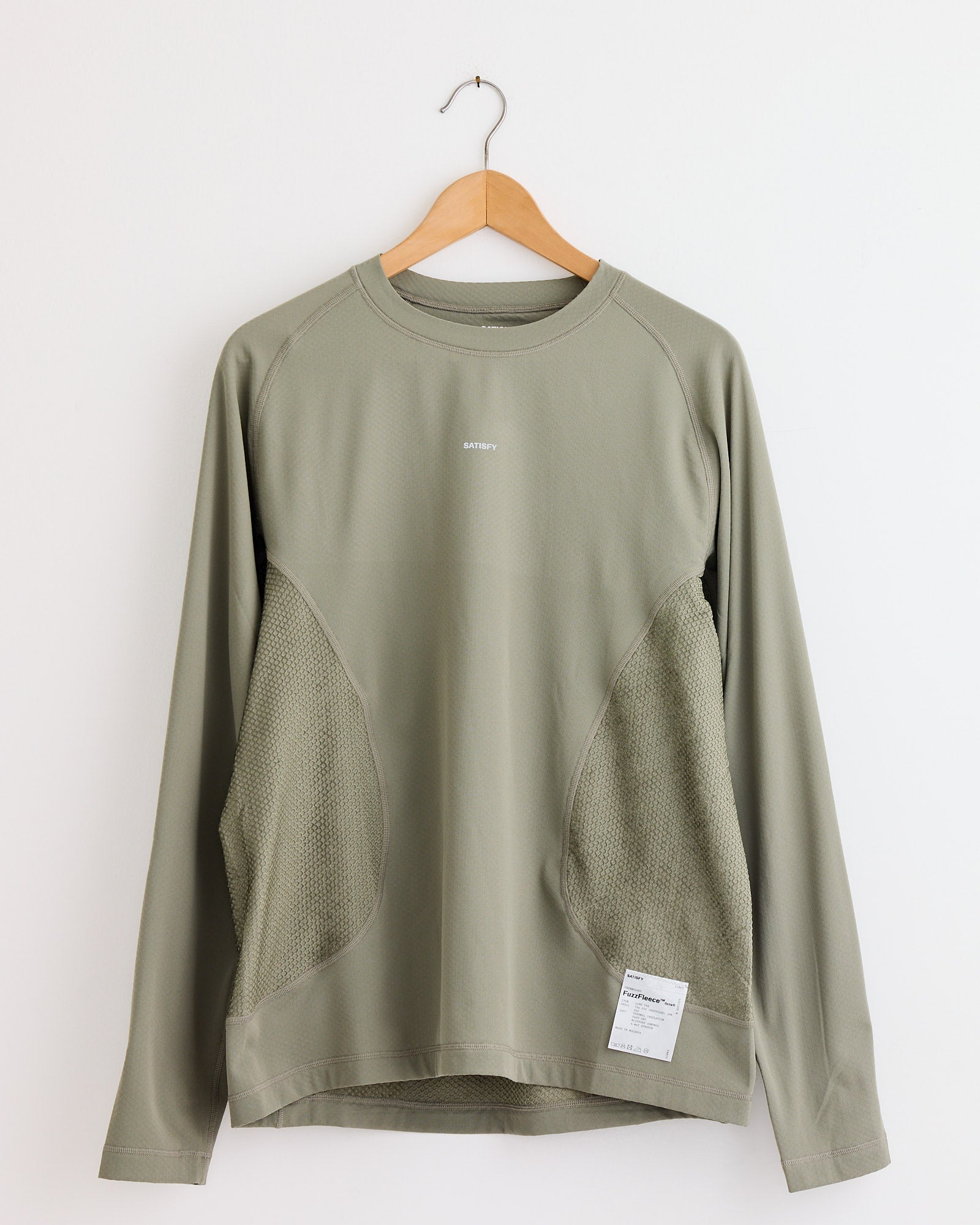 The Satisfy Running FuzzFleece Octa L/S T-Shirt in Vetiver features mesh side panels, a small white label at the hem, and a flattering fit. Displayed on a wooden hanger against a white wall. Chat online for sizing help.