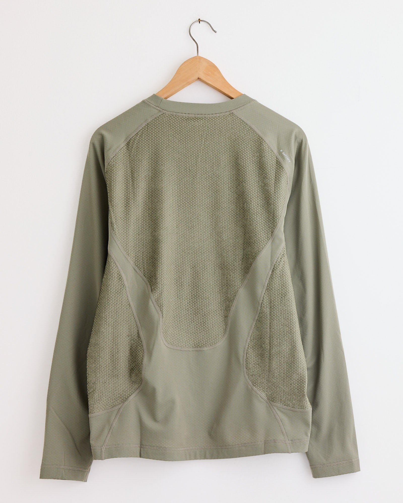 The Satisfy Running FuzzFleece Octa L/S T-Shirt in Vetiver, featuring mesh back panels, is displayed from the back on a wooden hanger against a white wall to highlight its fit and design.