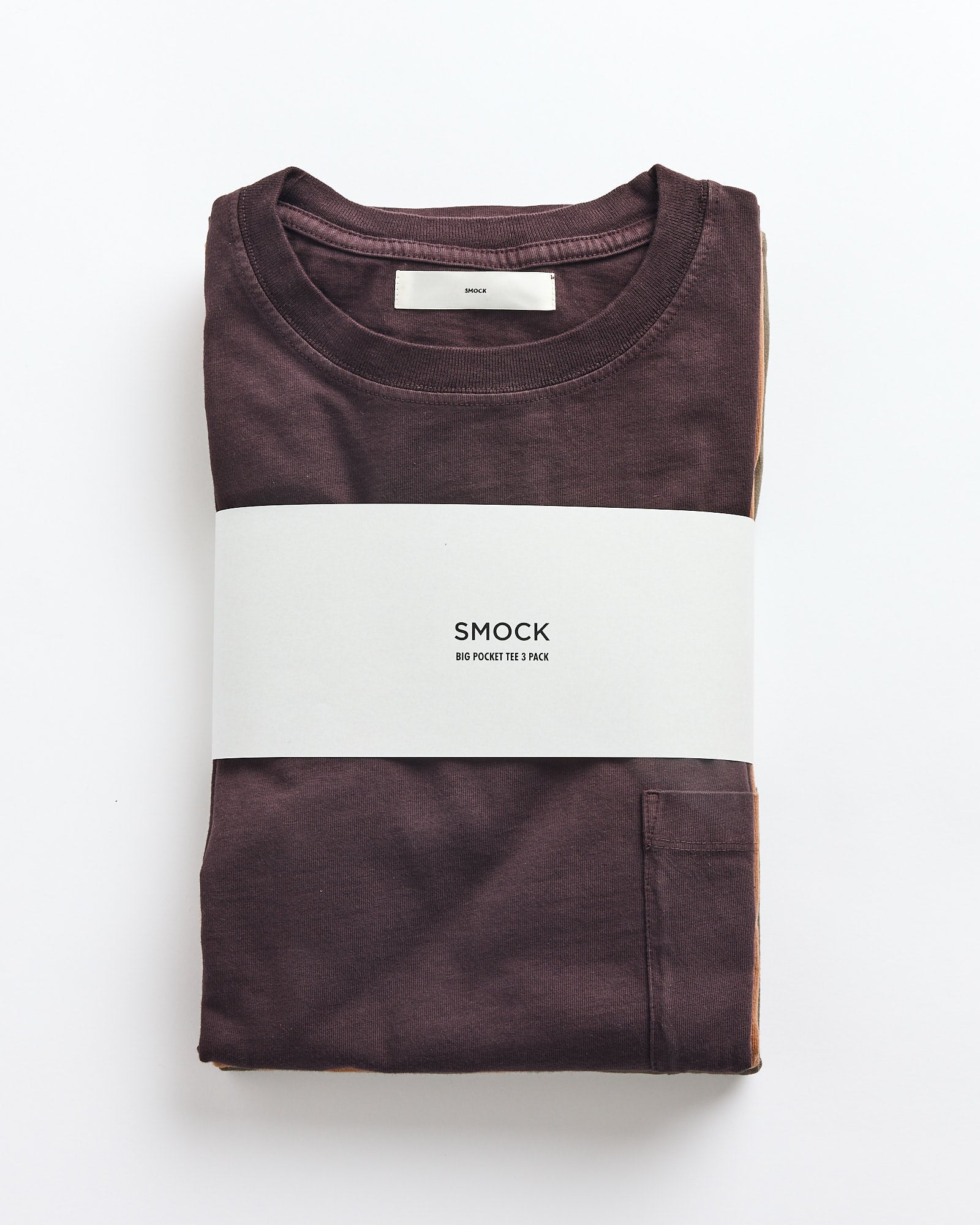 A folded SMOCK Bachelor Pack T-shirt in Caper/Port/Chai rests on a plain white background, featuring a white label with the brand name and small text wrapped around it. The tee has a visible chest pocket.