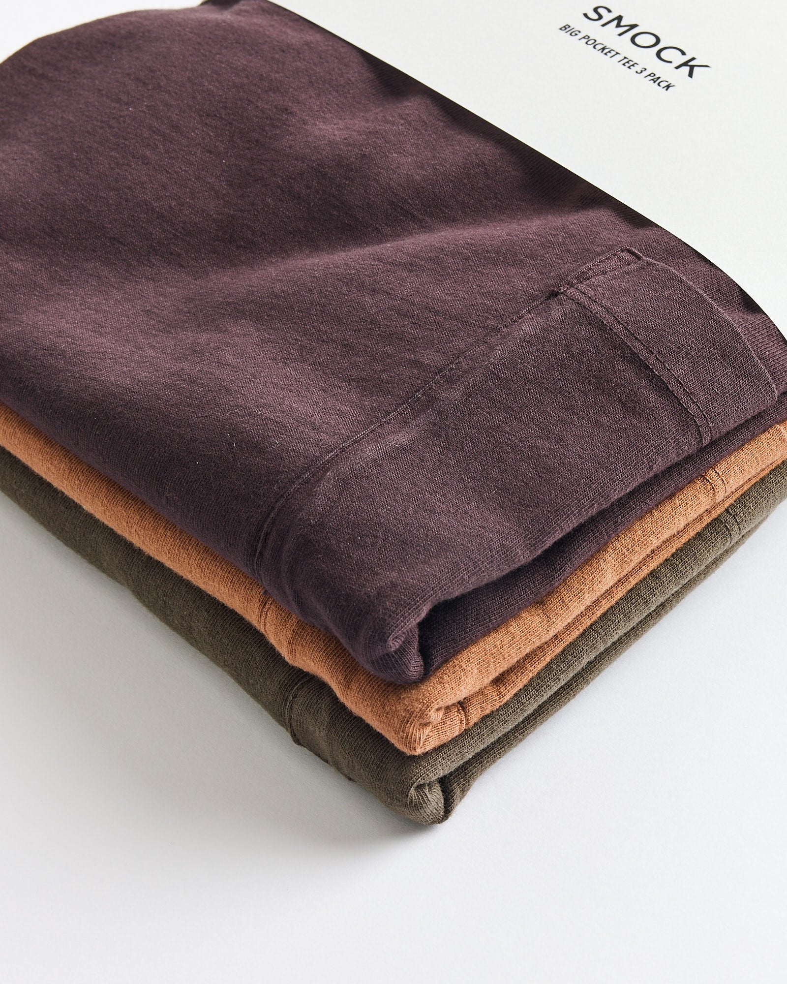 Three folded SMOCK Bachelor Pack tees in caper, port, and chai, neatly stacked with a white label reading SMOCK and Bachelor Pack. Features a boxy silhouette for a modern look.
