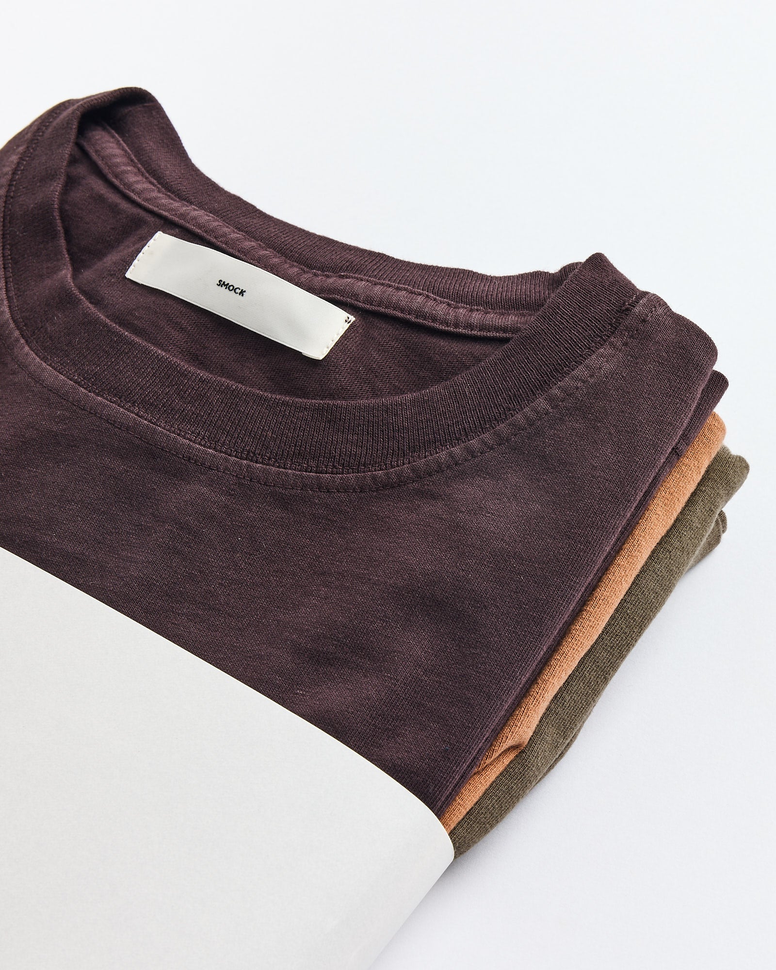 The SMOCK Bachelor Pack features three neatly folded combed cotton t-shirts in Caper, Port, and Chai hues with a visible white tag on top, displayed on a white surface for a refined, boxy silhouette.