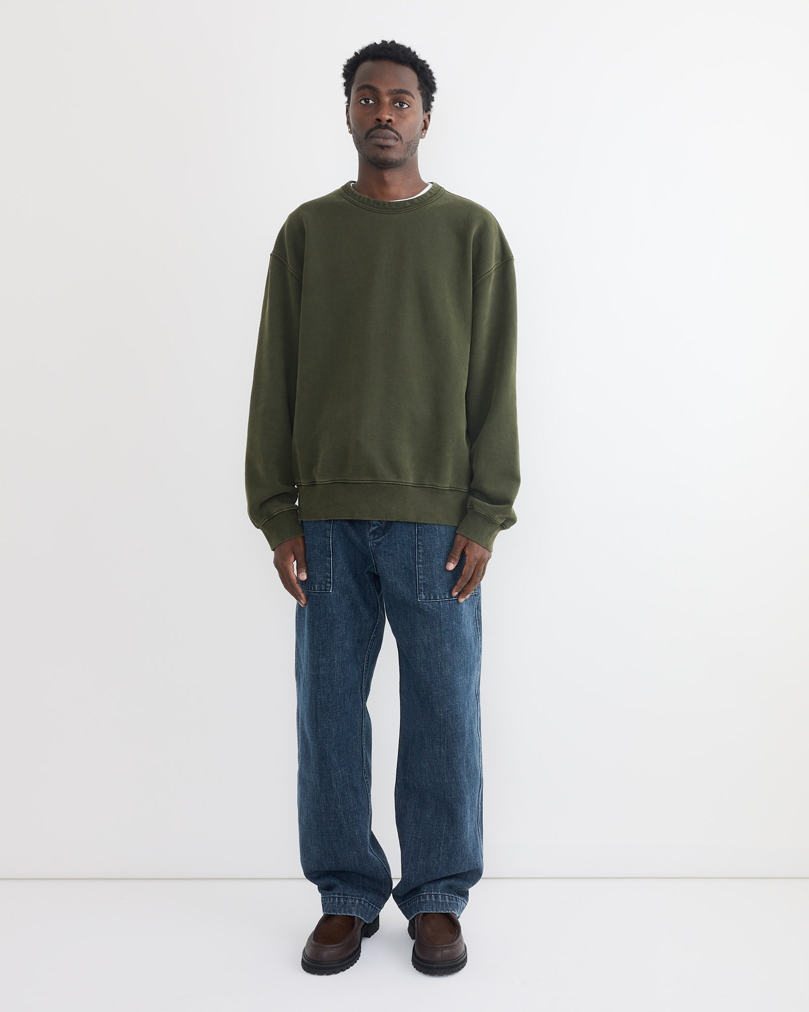 A man faces forward against a plain white background, wearing a loose Dolphy Crew in 22oz French Terry Green by SMOCK, paired with baggy blue jeans and dark brown shoes.