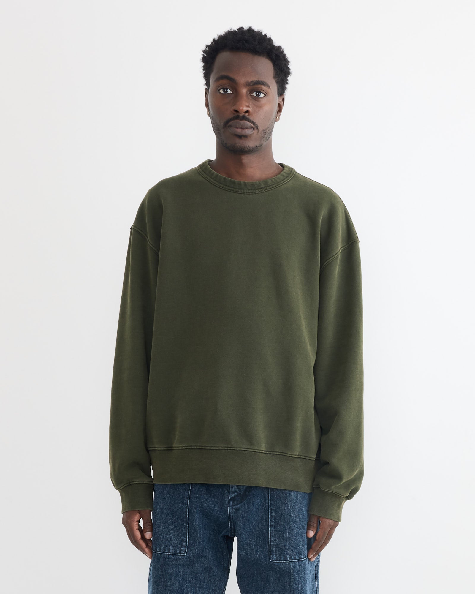 A man with short, curly hair and a neutral expression wears blue jeans and the SMOCK Dolphy Crew in 22oz French Terry Green, standing facing forward against a plain white background.