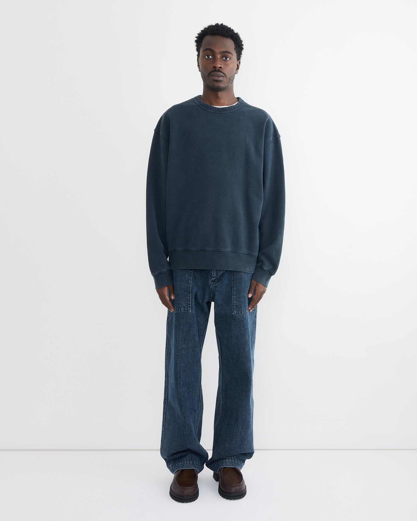 A man stands against a plain white background wearing the SMOCK Dolphy Crew in 22oz French Terry Navy, paired with baggy blue jeans and brown shoes. He looks directly at the camera with a neutral expression.