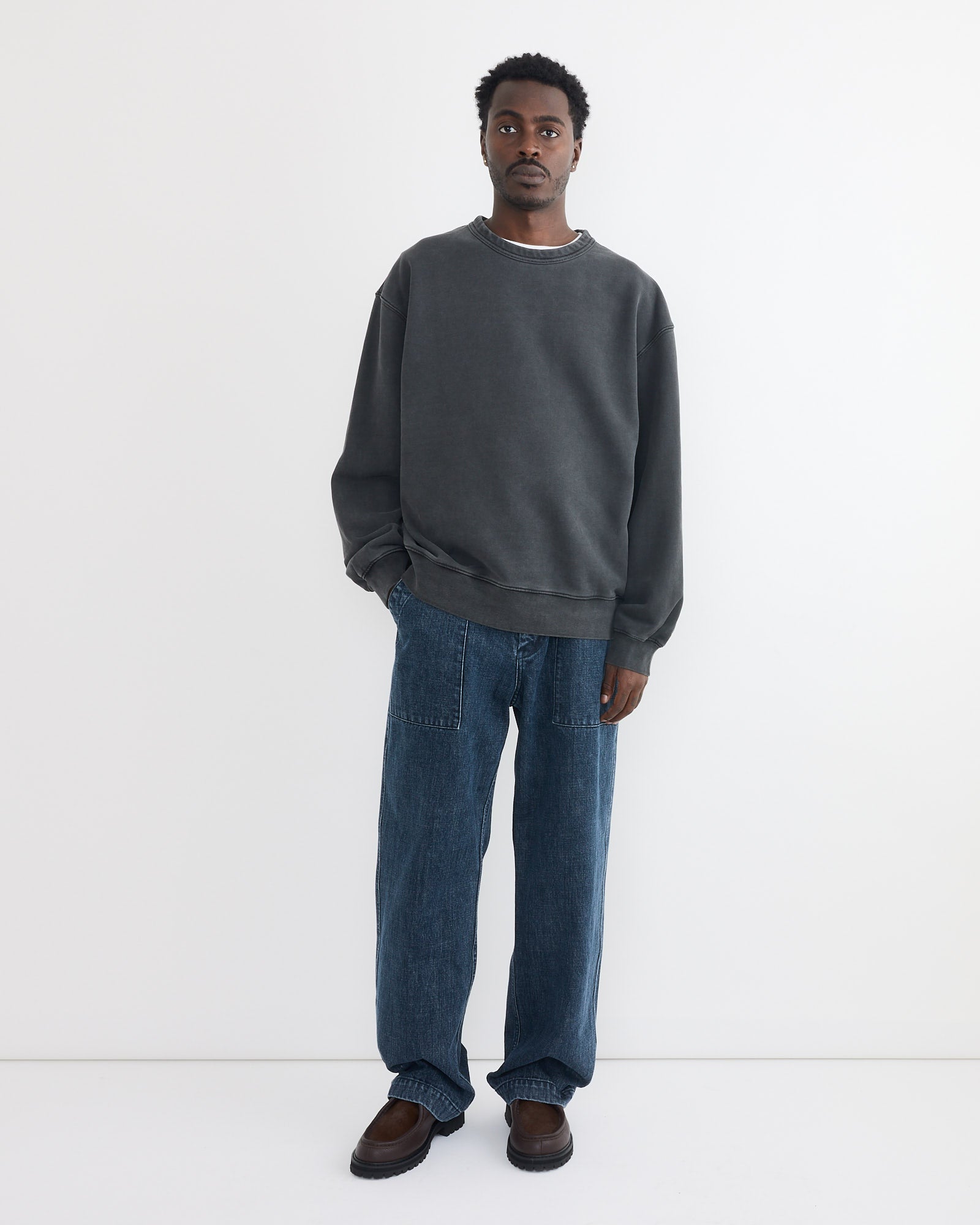 A man with short curly hair stands against a white background, wearing the SMOCK Dolphy Crew in 22oz French Terry Grey, paired with loose blue jeans and brown shoes. He has a neutral expression.