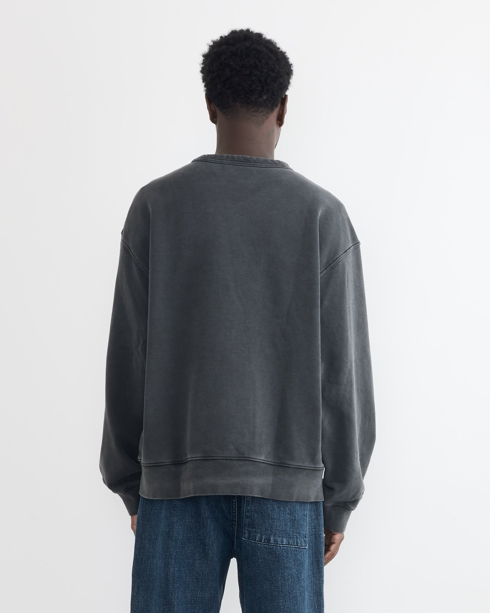 A person with short curly hair, seen from behind, wears a loose-fitting, faded dark gray SMOCK Dolphy Crew in 22oz French Terry Grey and blue jeans against a plain white background. Made in Los Angeles.