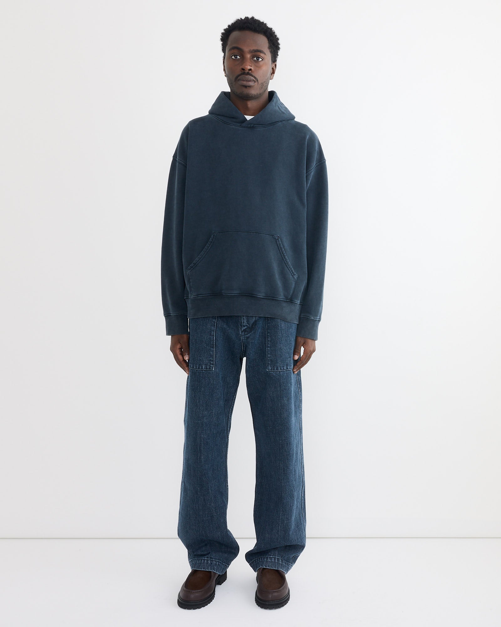 A person stands facing forward against a plain white background, wearing the SMOCK Ornette Hoodie in 22oz French Terry Navy, loose-fitting blue jeans, and brown shoes. Made in Los Angeles, they have a neutral expression with hands by their sides.