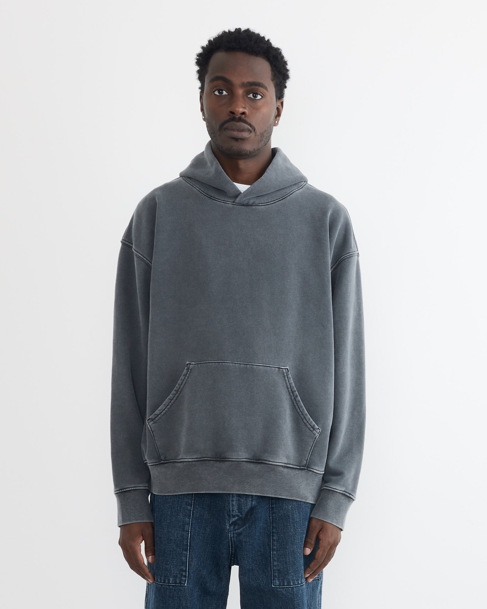 A man with short curly hair wears a loose-fitting SMOCK Ornette Hoodie in 22oz French Terry Grey, made in Los Angeles with a kangaroo pocket, and blue jeans, standing against a plain white background.