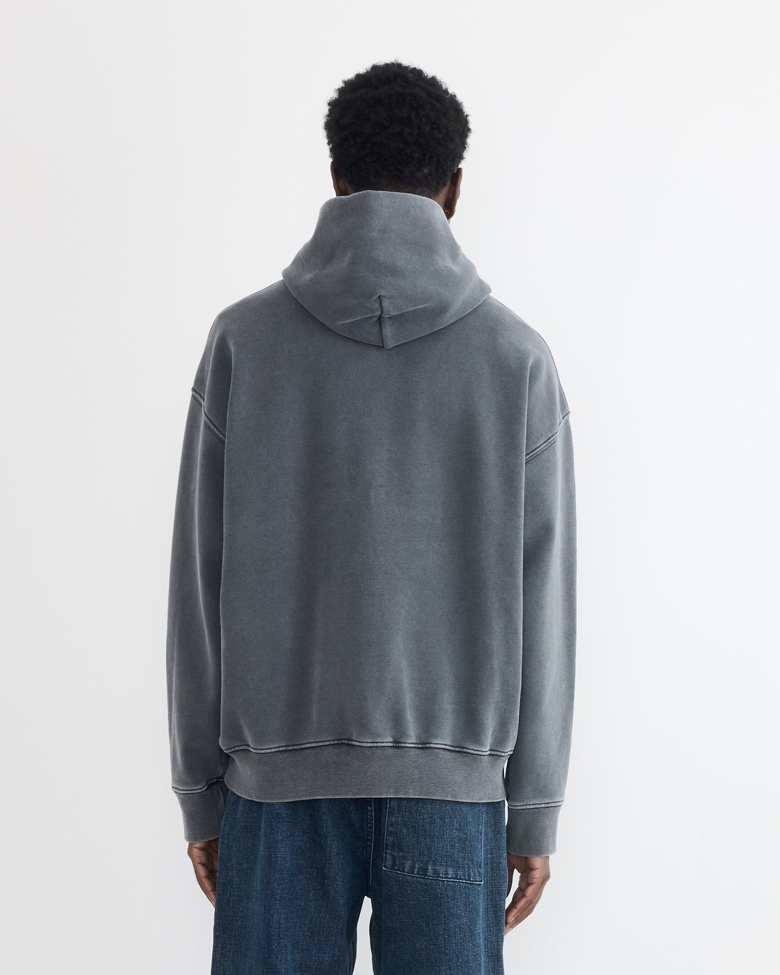 A person stands with their back to the camera, wearing the SMOCK Ornette Hoodie in 22oz French Terry Grey and blue jeans. The hoodie’s faded grey hood is up. Made in Los Angeles, shown against a plain white background.