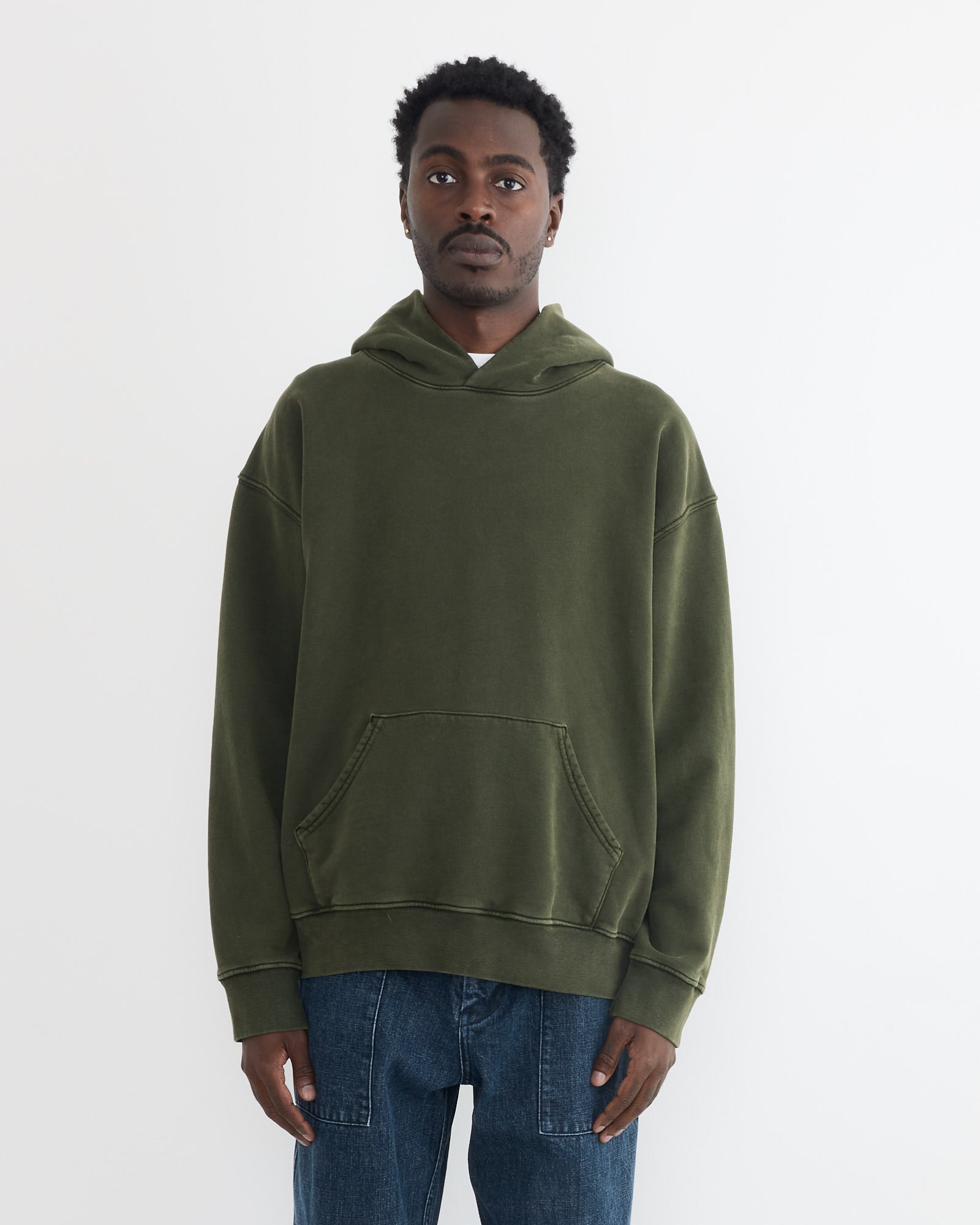 A person stands against a white background wearing the SMOCK Ornette Hoodie in 22oz French Terry Green and blue jeans, hands relaxed by their sides. Made in Los Angeles.