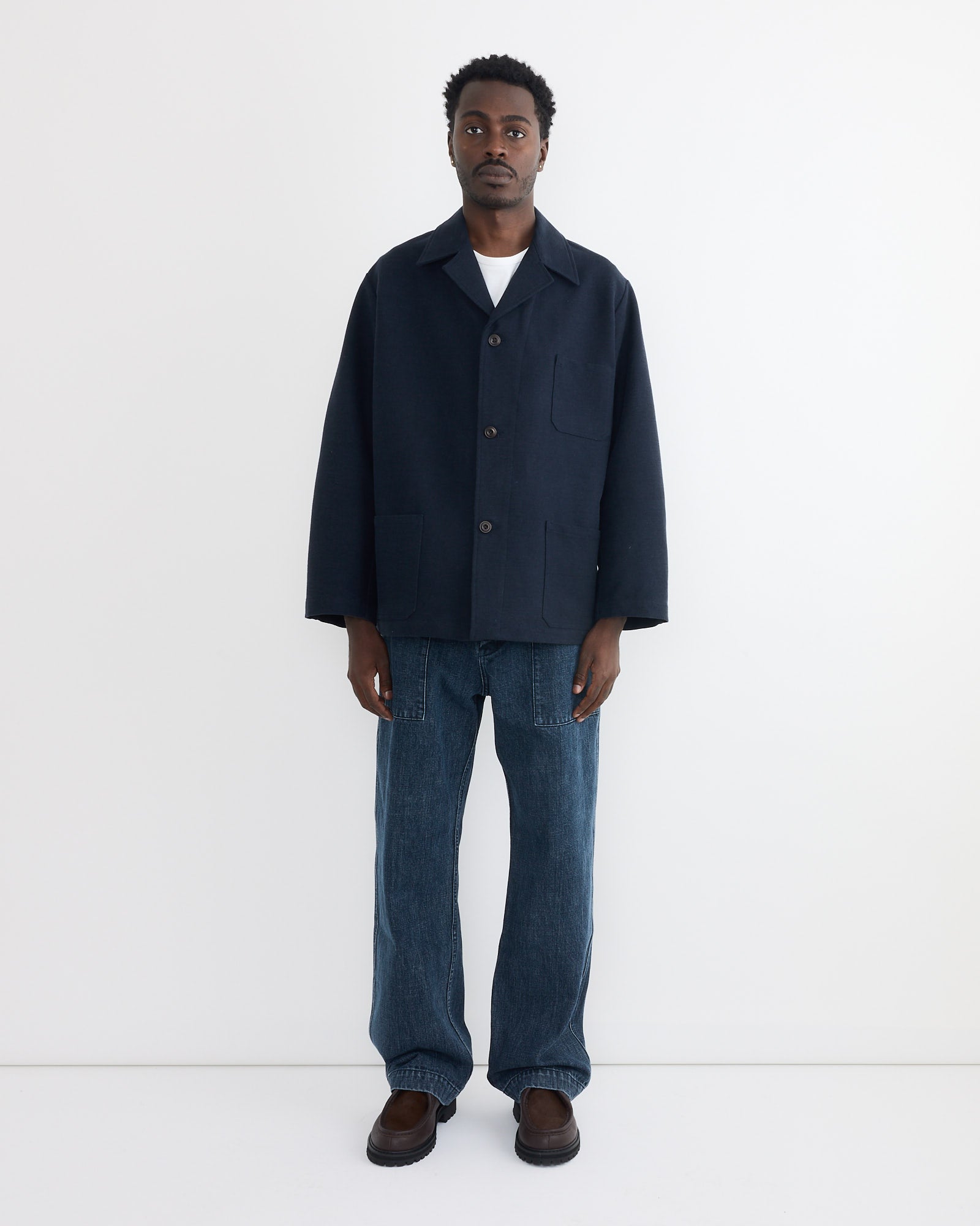 A man stands against a white background wearing SMOCK's Toyon Pant in Denim—relaxed fit, deadstock Japanese denim—paired with a navy button-up jacket, white shirt, and brown shoes. He faces forward with his arms at his sides.