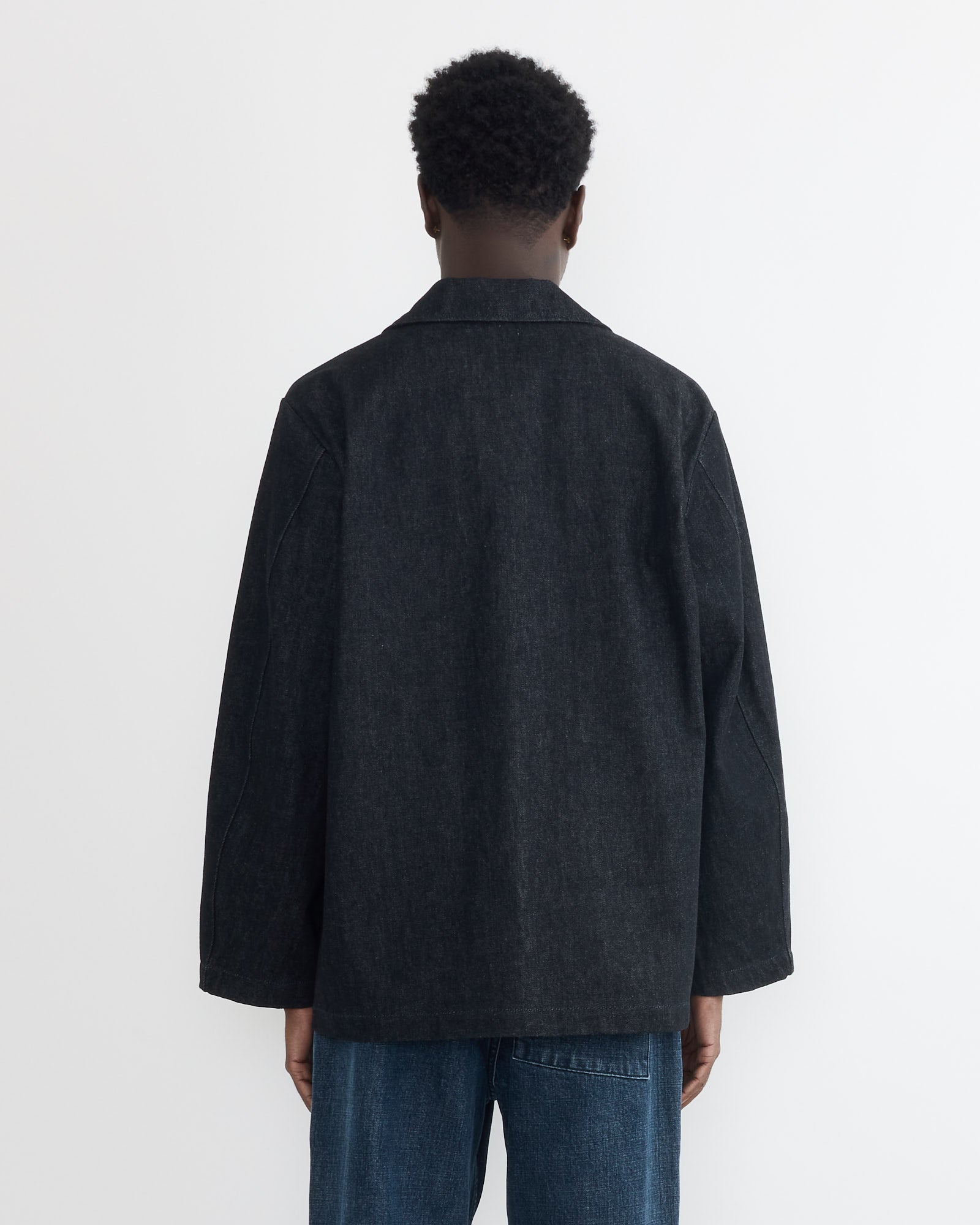 A person with short curly hair, facing away from the camera, wears the SMOCK Vannier Jacket in Denim and blue jeans against a plain white background.