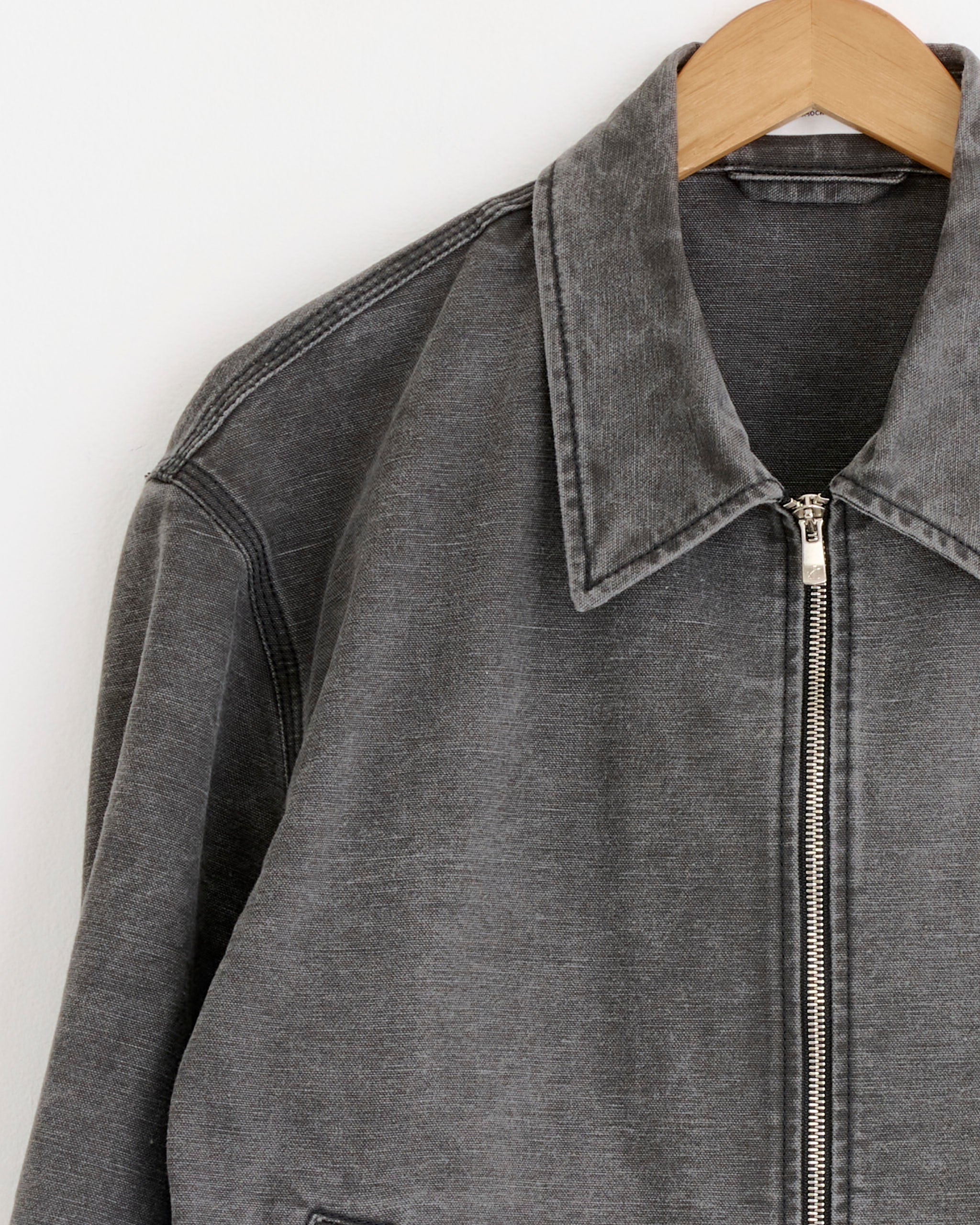 A close-up of the SMOCK Triple Stitch Hydra Jacket in Washed Pigment Dyed Cotton Grey on a wooden hanger against a white wall, highlighting the collar, zipper, and sleeve—ideal for assessing fit and sizing before purchase.