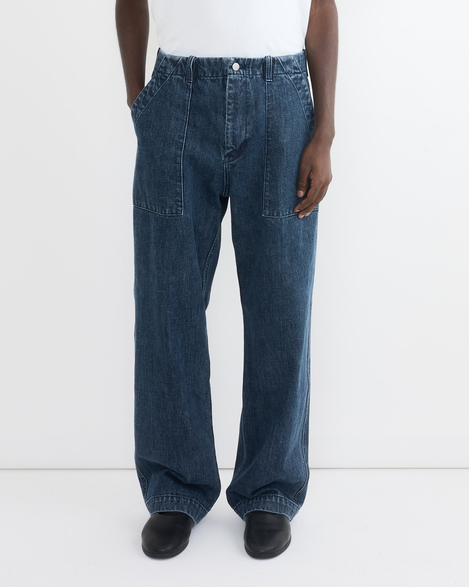 A person stands against a plain white background wearing SMOCK’s Toyon Pant in Denim, relaxed fit and crafted from deadstock Japanese denim, paired with a tucked-in white shirt and black shoes.