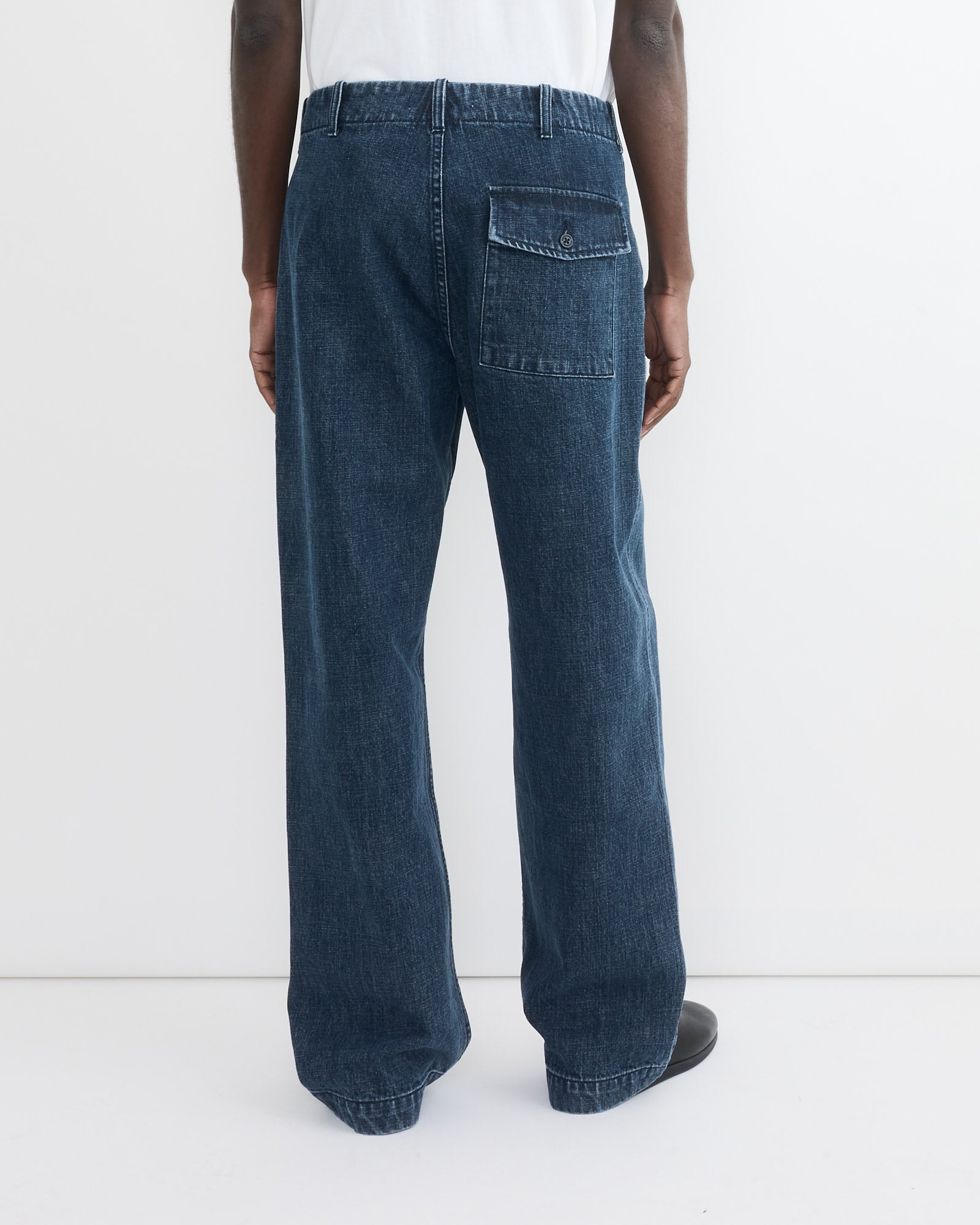 A person faces away, wearing SMOCK's Toyon Pant in Denim—relaxed fit, deadstock Japanese denim with an oversized buttoned back pocket—paired with a white shirt and black shoes against a plain white background.