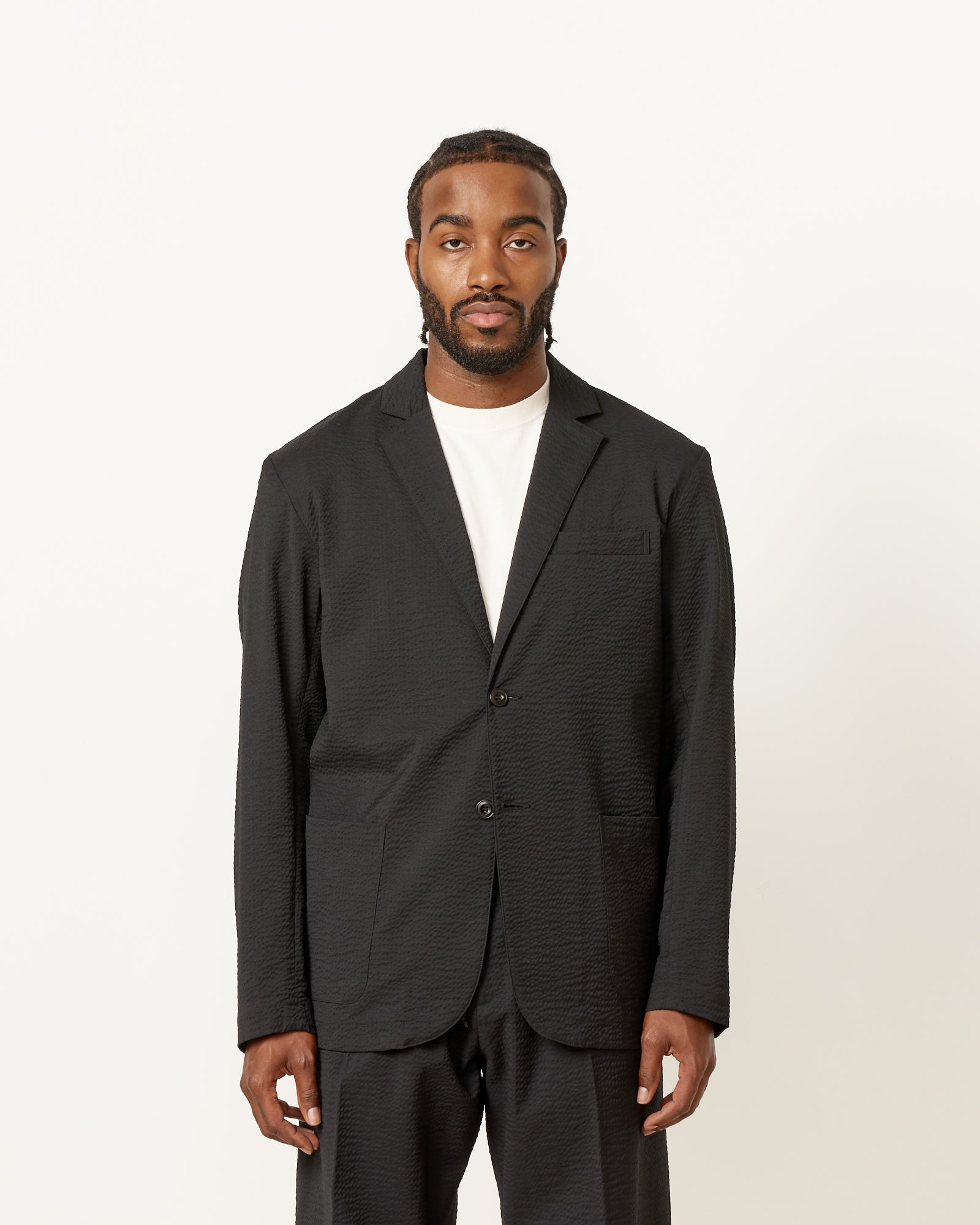 A man with short hair and a beard faces the camera, wearing the SMOCK Blazer Super 120s Wool Seersucker in Black over a white T-shirt and matching black pants, styled by Mohawk General Store against a plain white background.