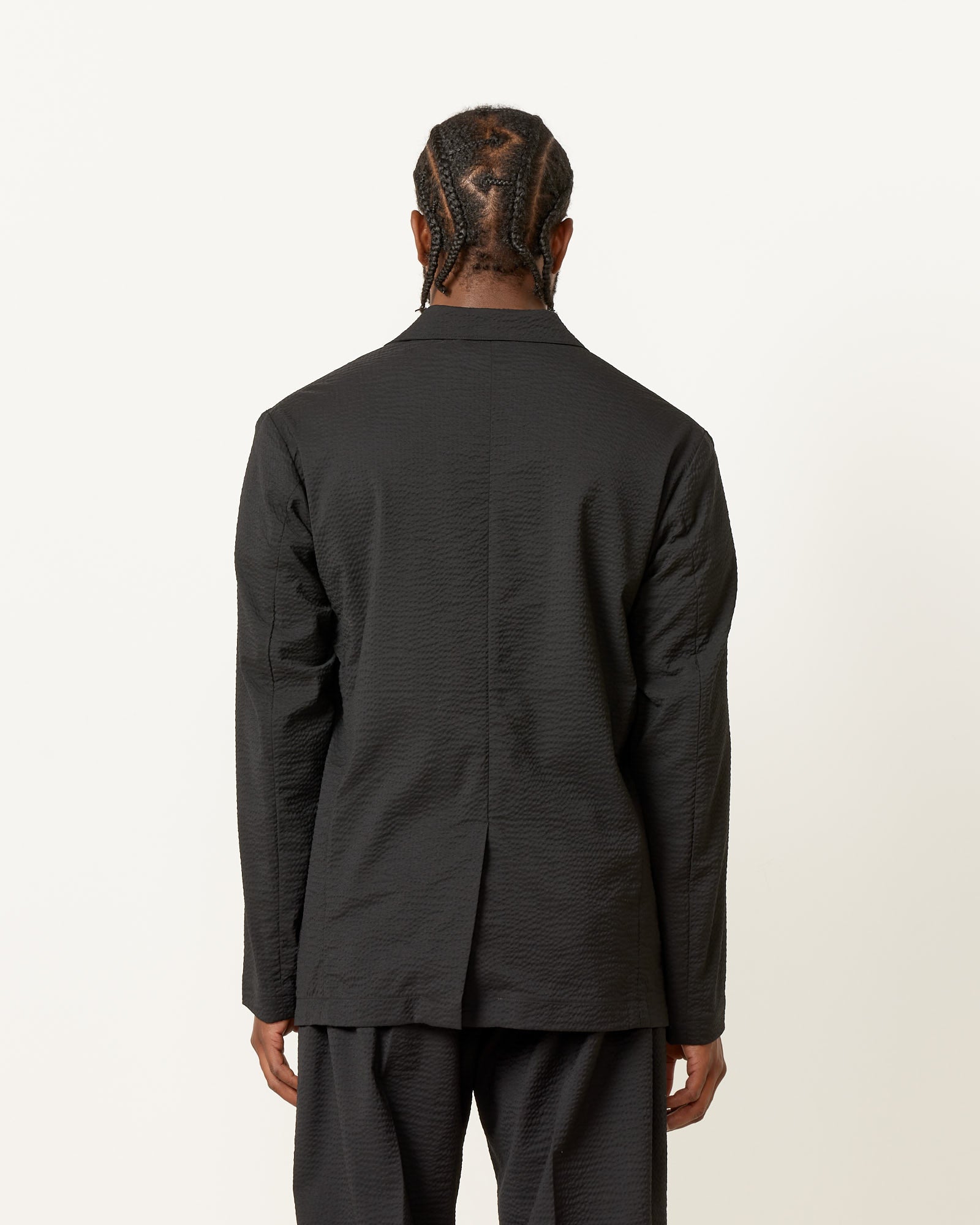 A person with braided hair faces away from the camera, wearing a textured black SMOCK Blazer Super 120s Wool Seersucker in Black, from Mohawk General Store, set against a plain white background.