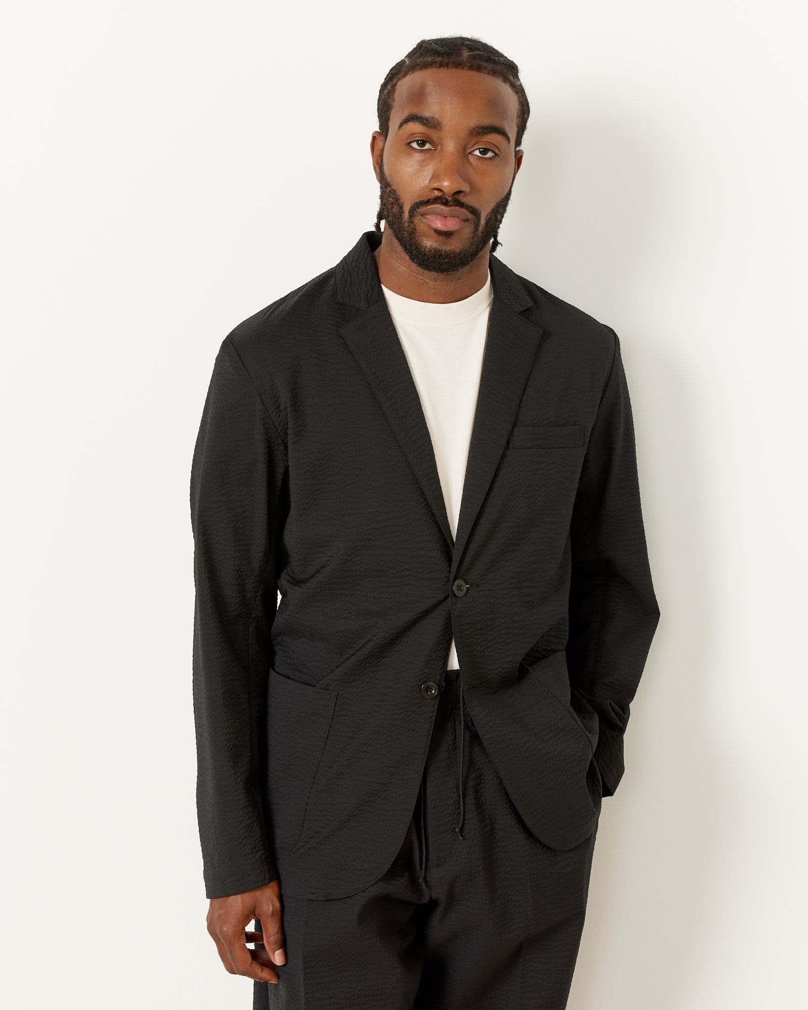 A man with short braids and a beard wears the SMOCK Blazer Super 120s Wool Seersucker in Black over a white t-shirt, standing against a plain white background with one hand in his pocket.