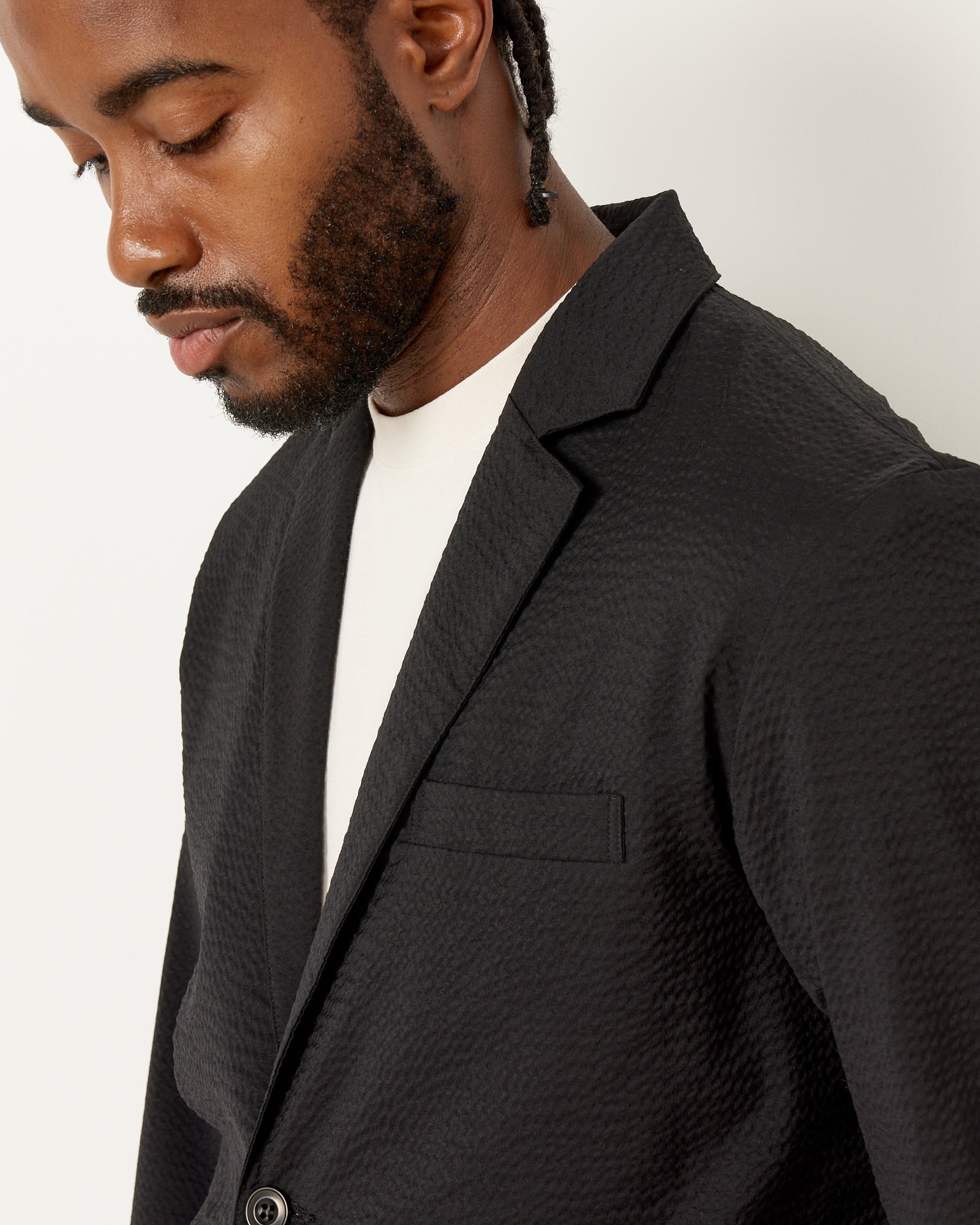 A man with a short beard and braided hair wears the SMOCK Blazer Super 120s Wool Seersucker in Black over a white shirt, looking downward against a plain white background—a refined look from Mohawk General Store.