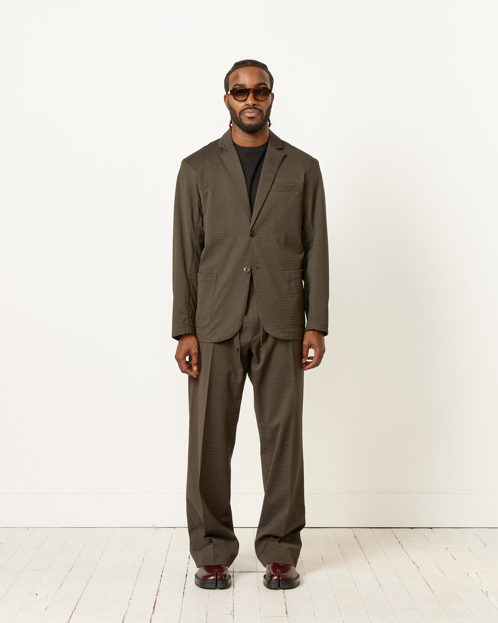 A man with short hair and a beard stands against a white wall, wearing an olive SMOCK Blazer Super 120s Wool Seersucker from Mohawk General Store, paired with a black shirt, brown shoes, and sunglasses. His hands are relaxed by his sides.