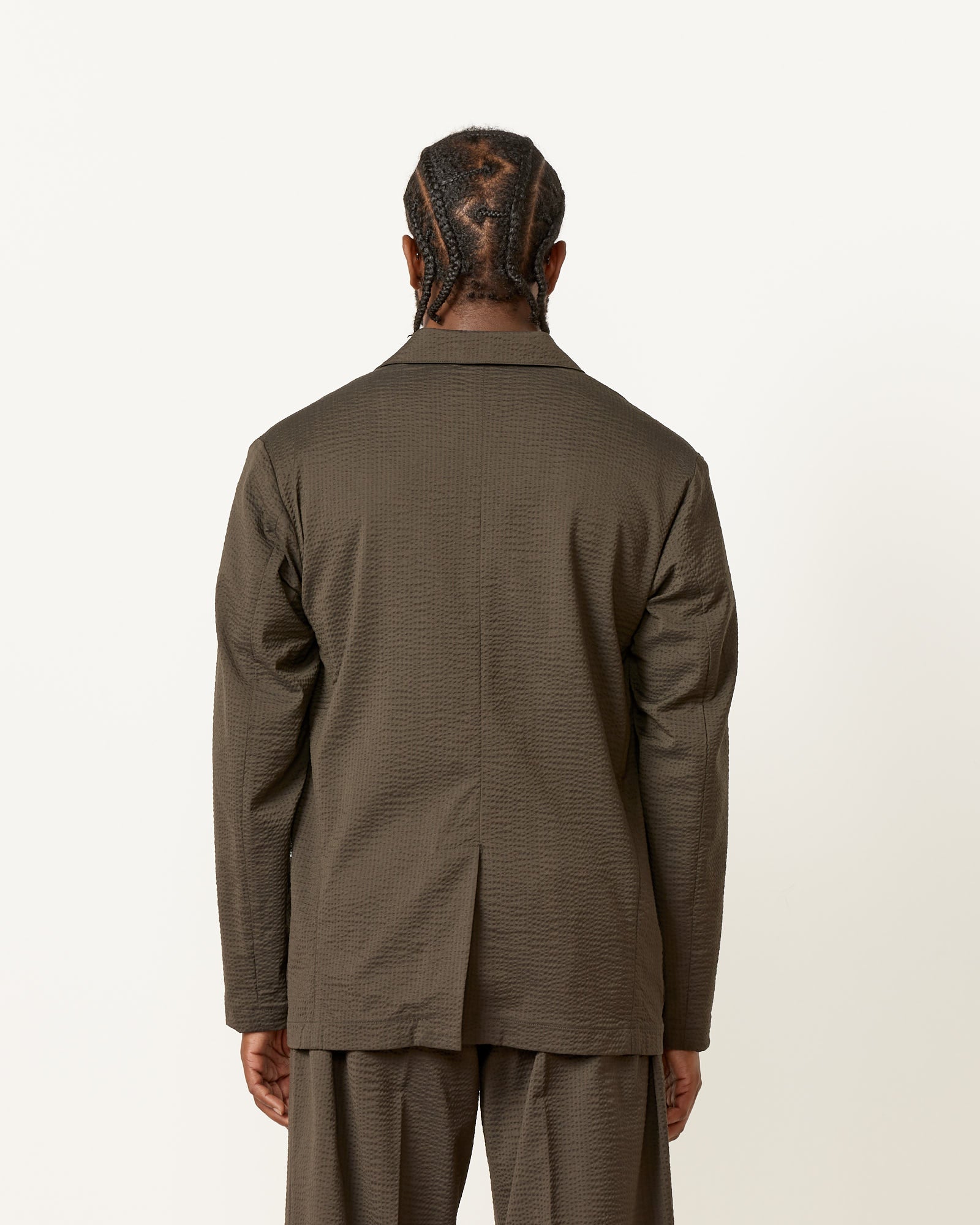 A person with braided hair, wearing the SMOCK Blazer Super 120s Wool Seersucker in Olive, stands with their back to the camera against a plain white background.